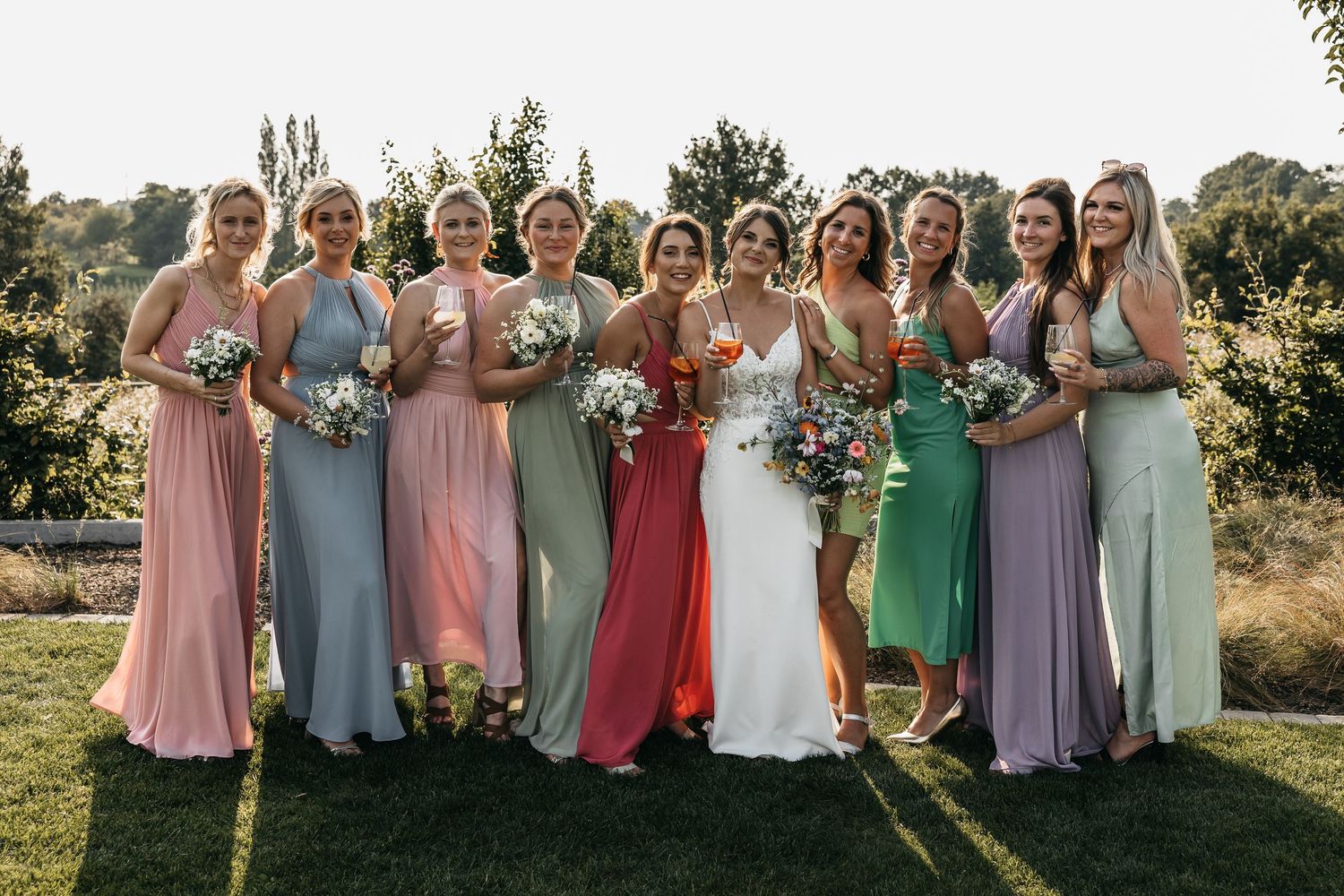 Bride poses with eight bridesmaids in colorful pastel gowns holding bouquets outdoors.