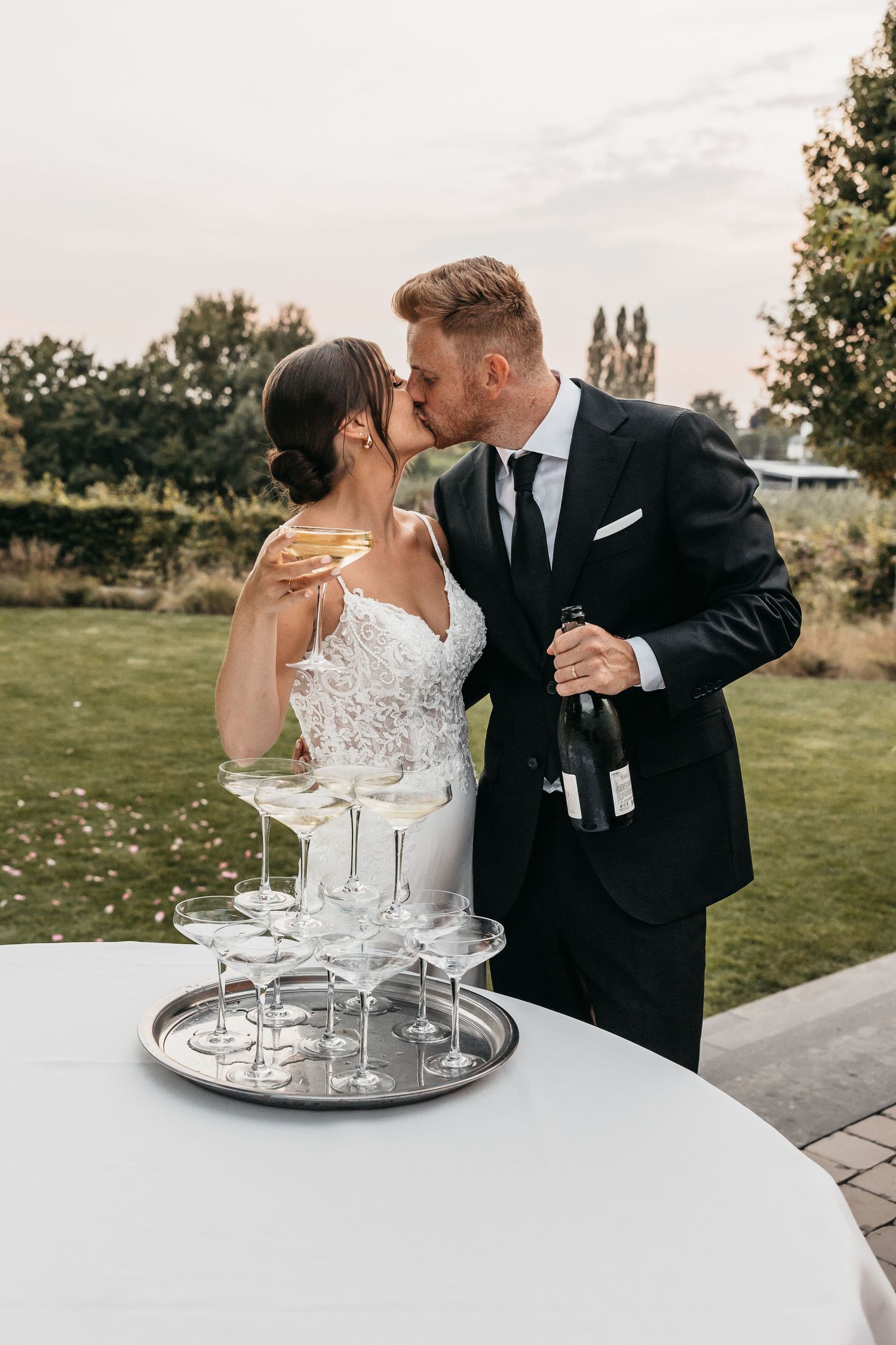 Bride and groom kissing beside champagne tower at elegant outdoor wedding reception.