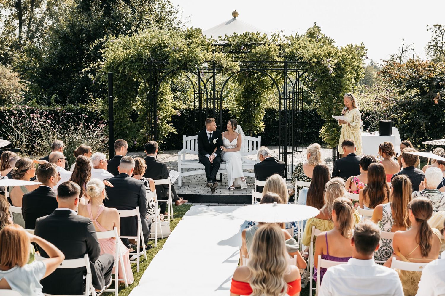 Wide view of outdoor wedding ceremony with guests seated in rows facing couple under garden pergola.