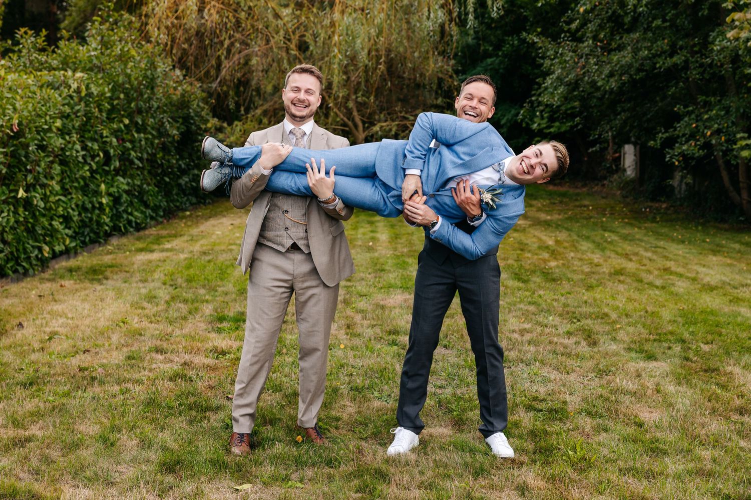 Two groomsmen playfully carry a third man horizontally while laughing outdoors on a green lawn.