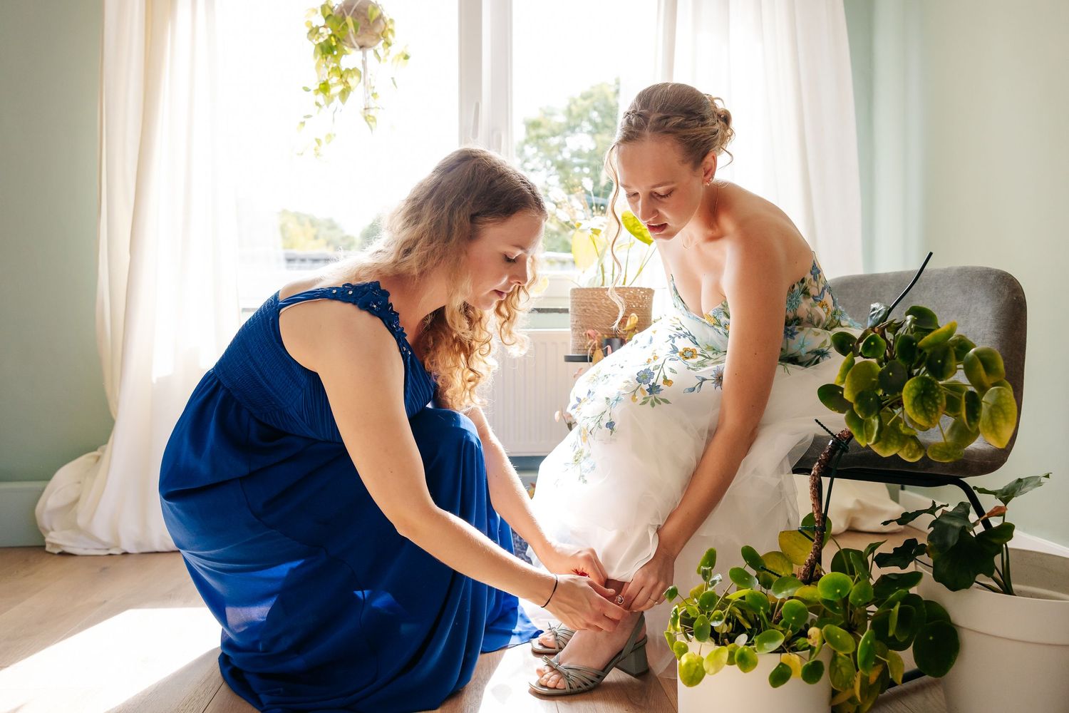 Bridesmaid in blue dress helping bride with sandal straps while bride smiles warmly.