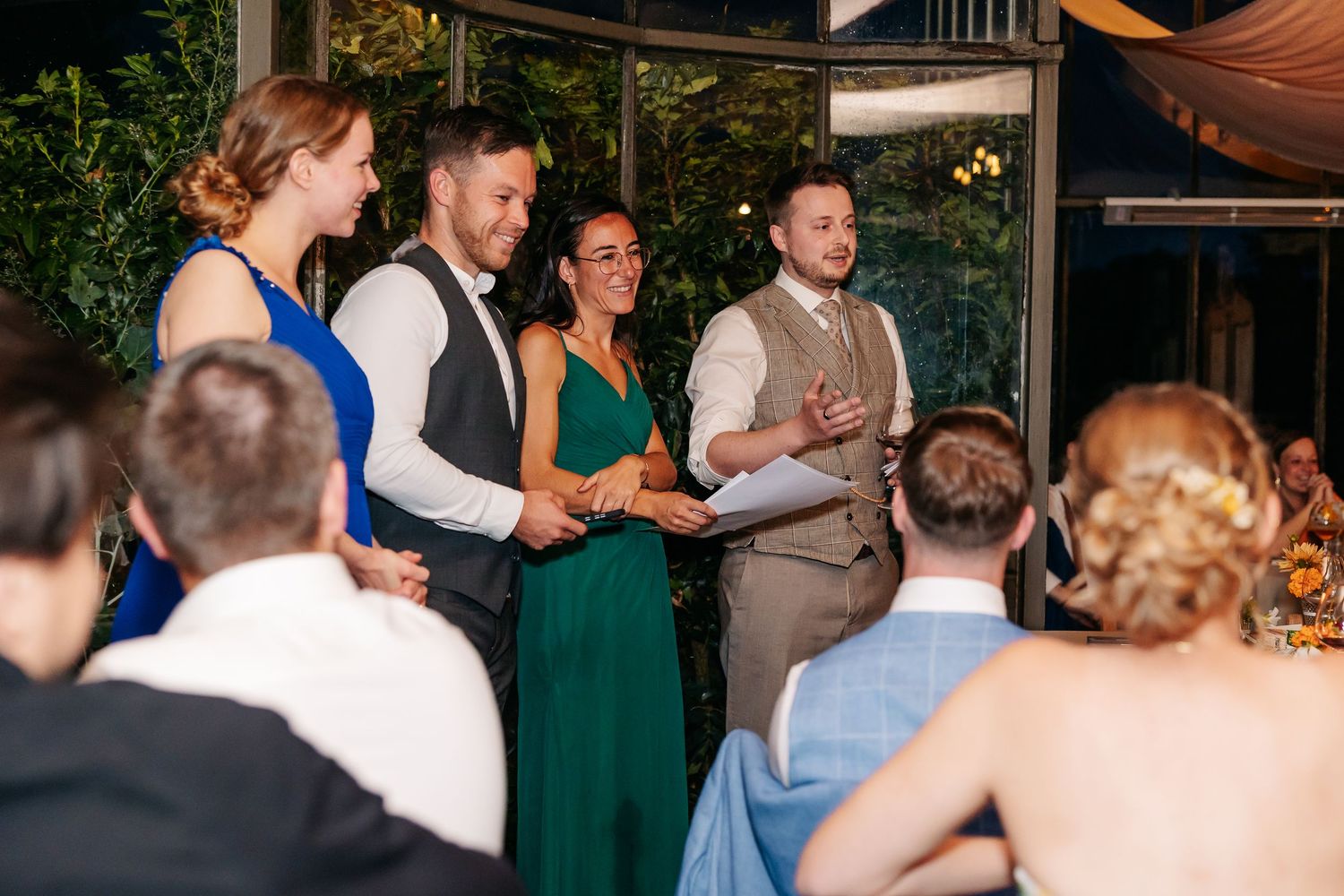 Wedding party members deliver a speech to seated guests at an elegant indoor reception venue.