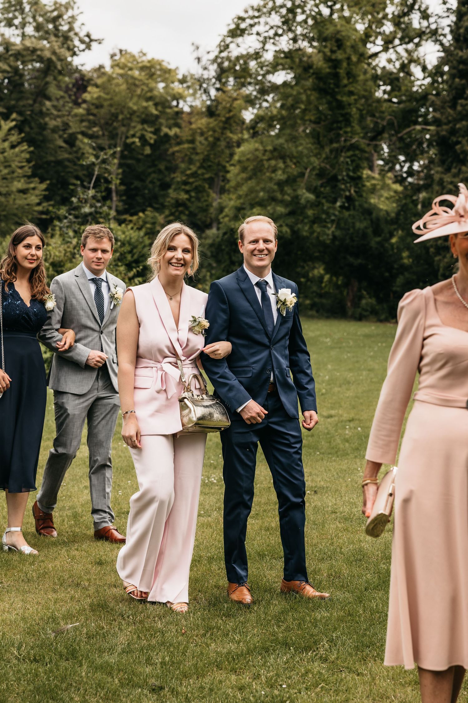 Elegantly dressed wedding guests walk across green lawn during outdoor summer wedding celebration.
