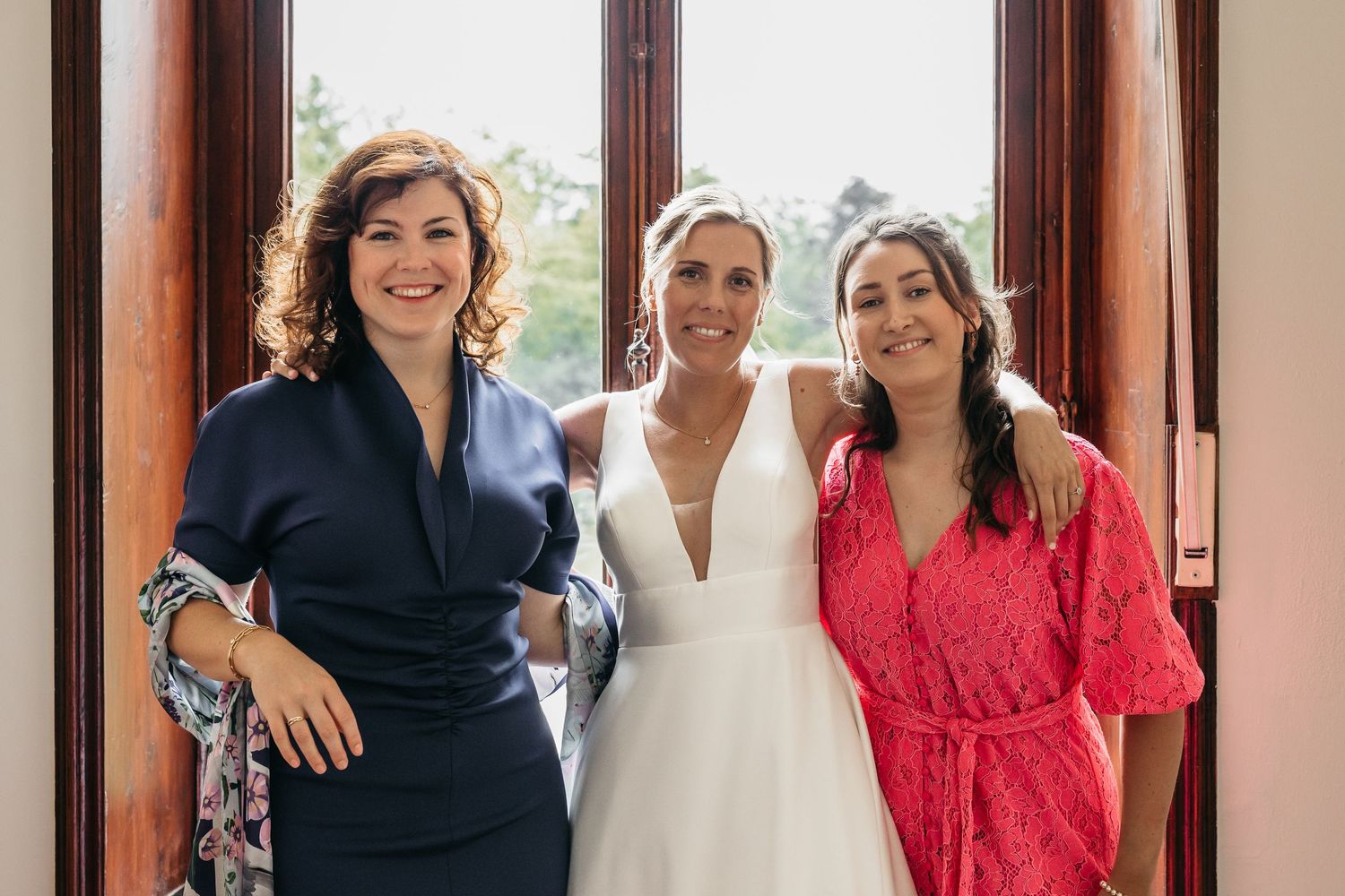 Bride in white gown posing with two smiling women in navy and red dresses at doorway.