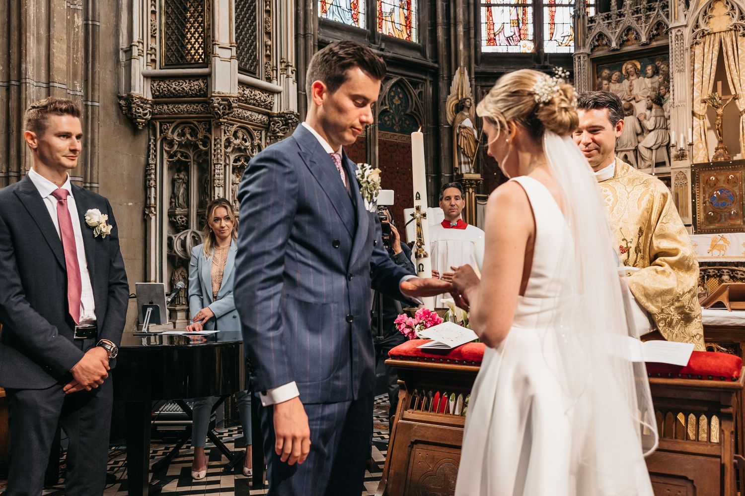 Groom in navy suit bows head during ring exchange at church wedding.