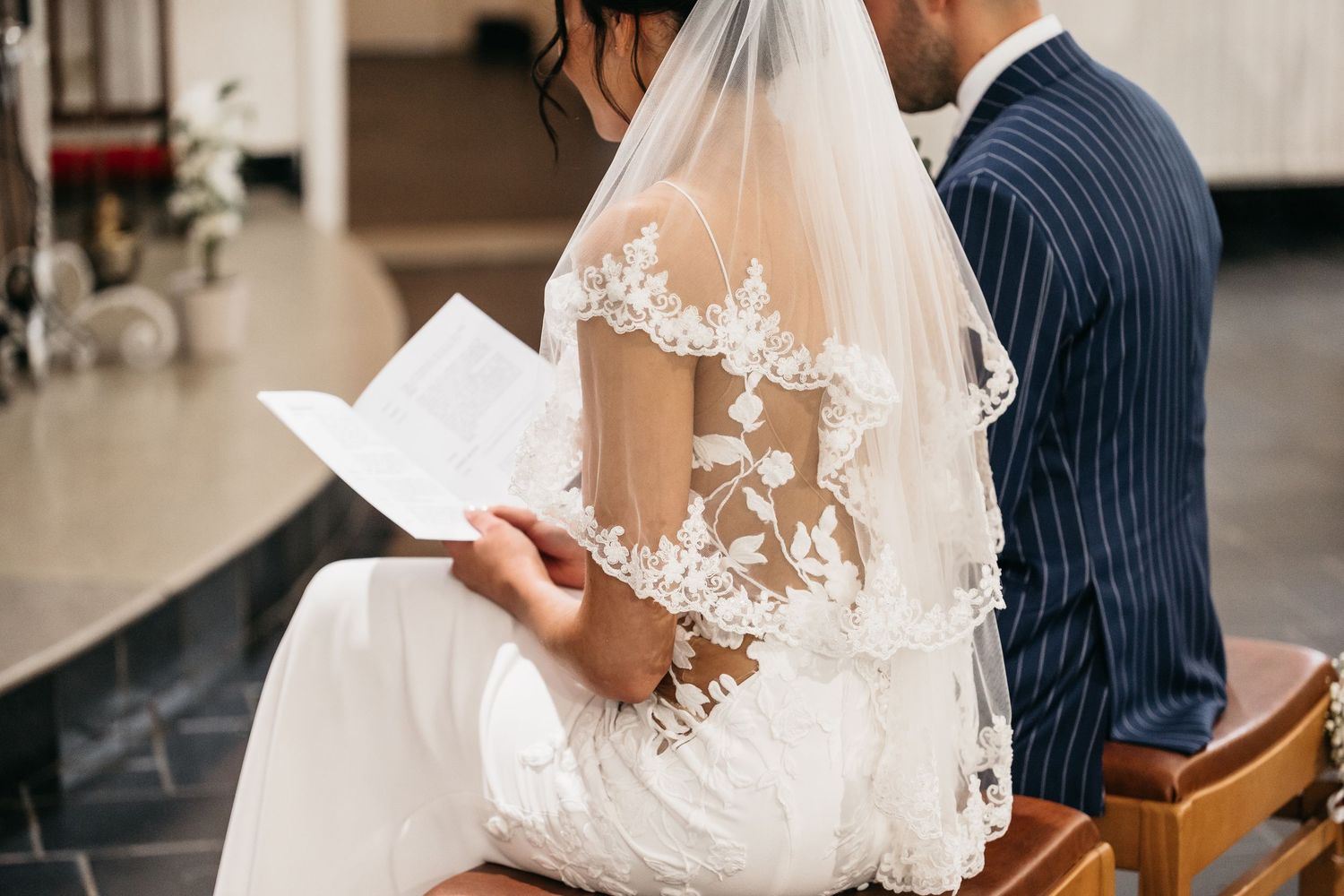 Bride in floral lace wedding dress reads ceremony program during church wedding.