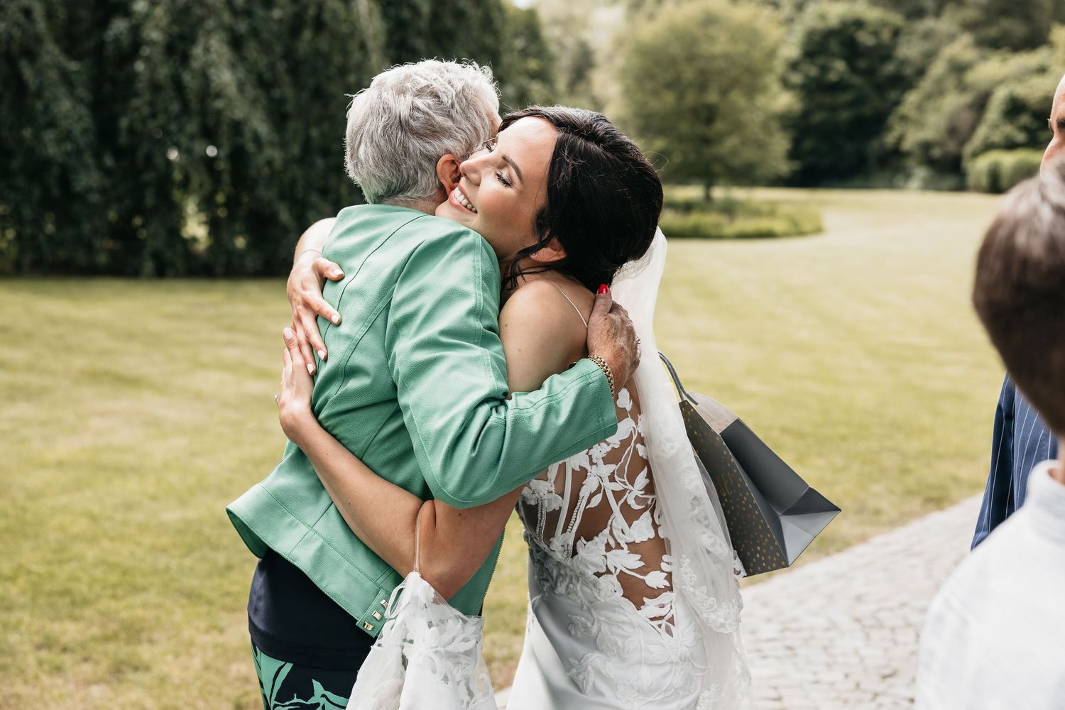 Bride in lace wedding gown sharing a warm embrace with an elderly woman in a green jacket outdoors.