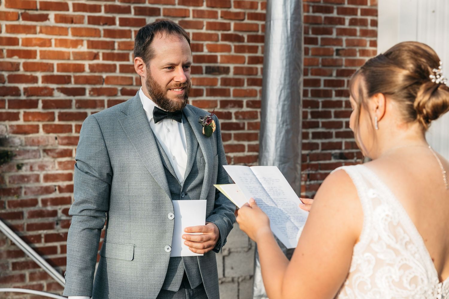 Groom in gray suit listens emotionally as bride reads wedding vows outside brick building during first look.