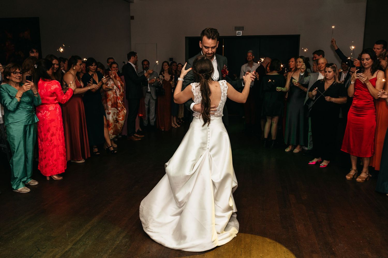 Groom dips bride in white satin gown during first dance as guests watch with sparklers.
