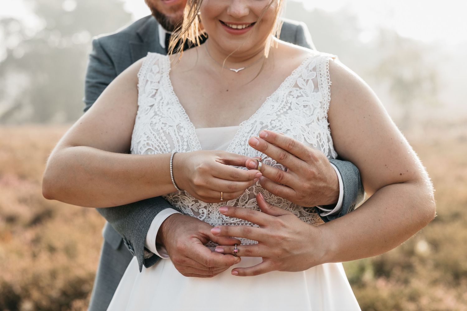 Groom embracing bride from behind, showing wedding rings and bridal jewelry in soft natural light.