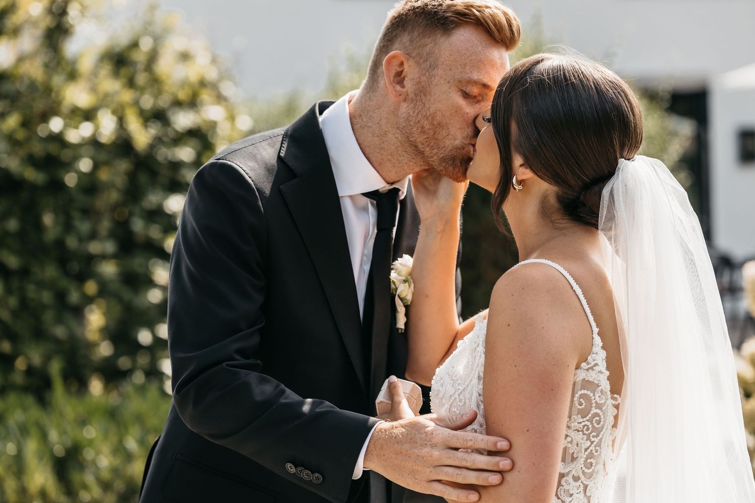 Groom in black suit kisses bride's forehead tenderly while embracing her in sunny outdoor garden setting.