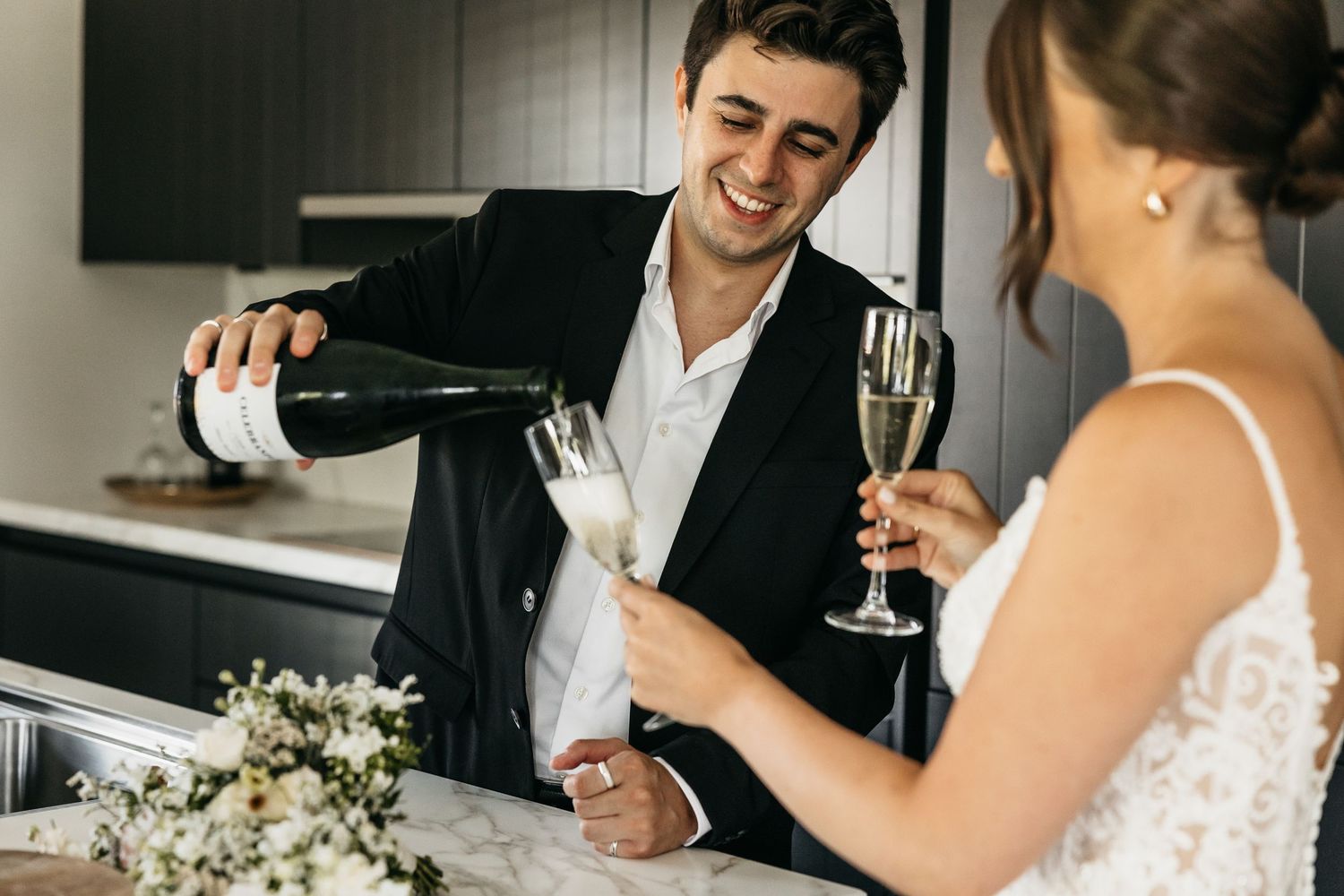 Smiling groom pouring champagne for bride at marble kitchen counter with flowers.