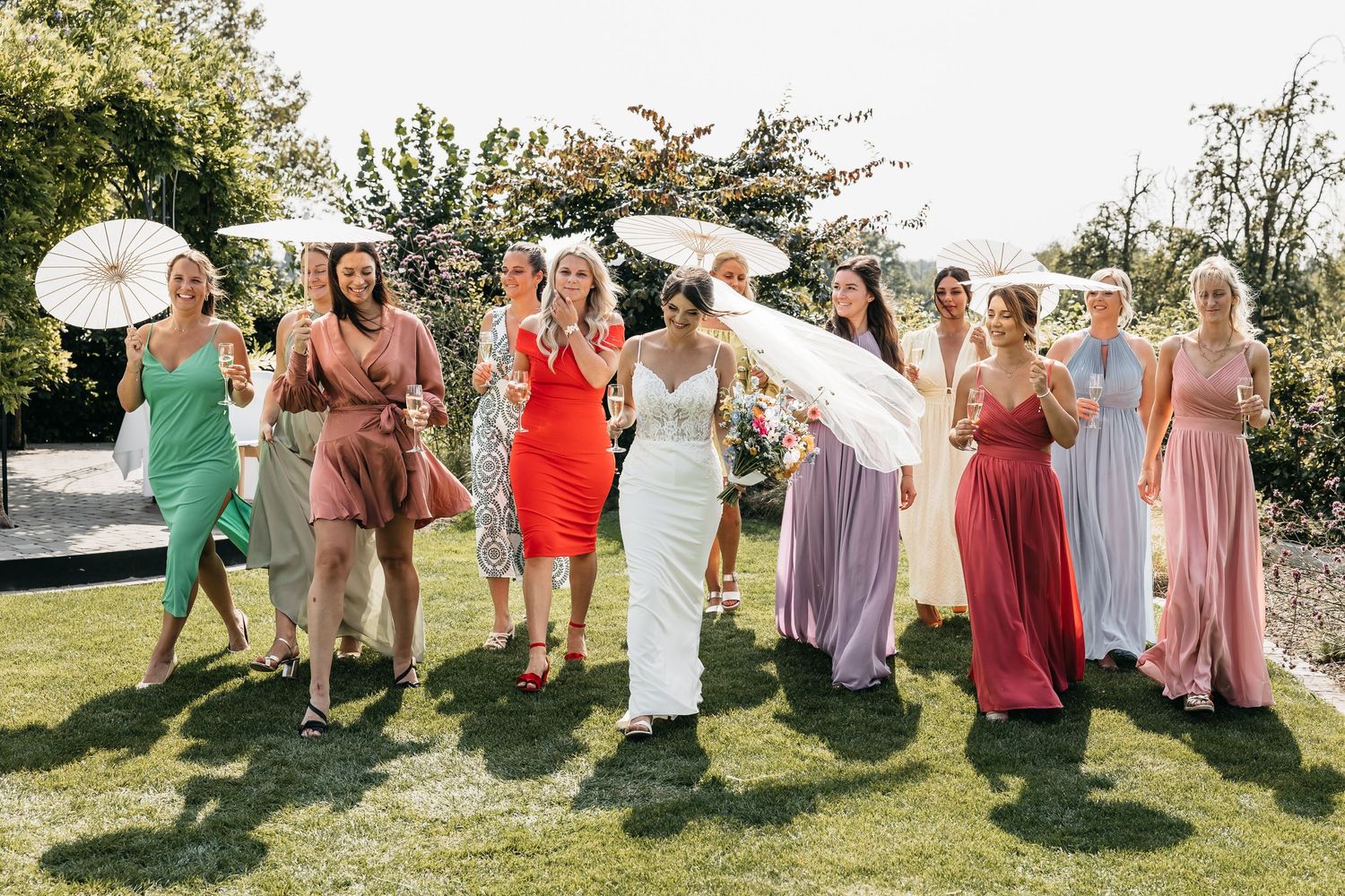 Bride in white gown laughing with bridesmaids in colorful dresses holding parasols at outdoor wedding.