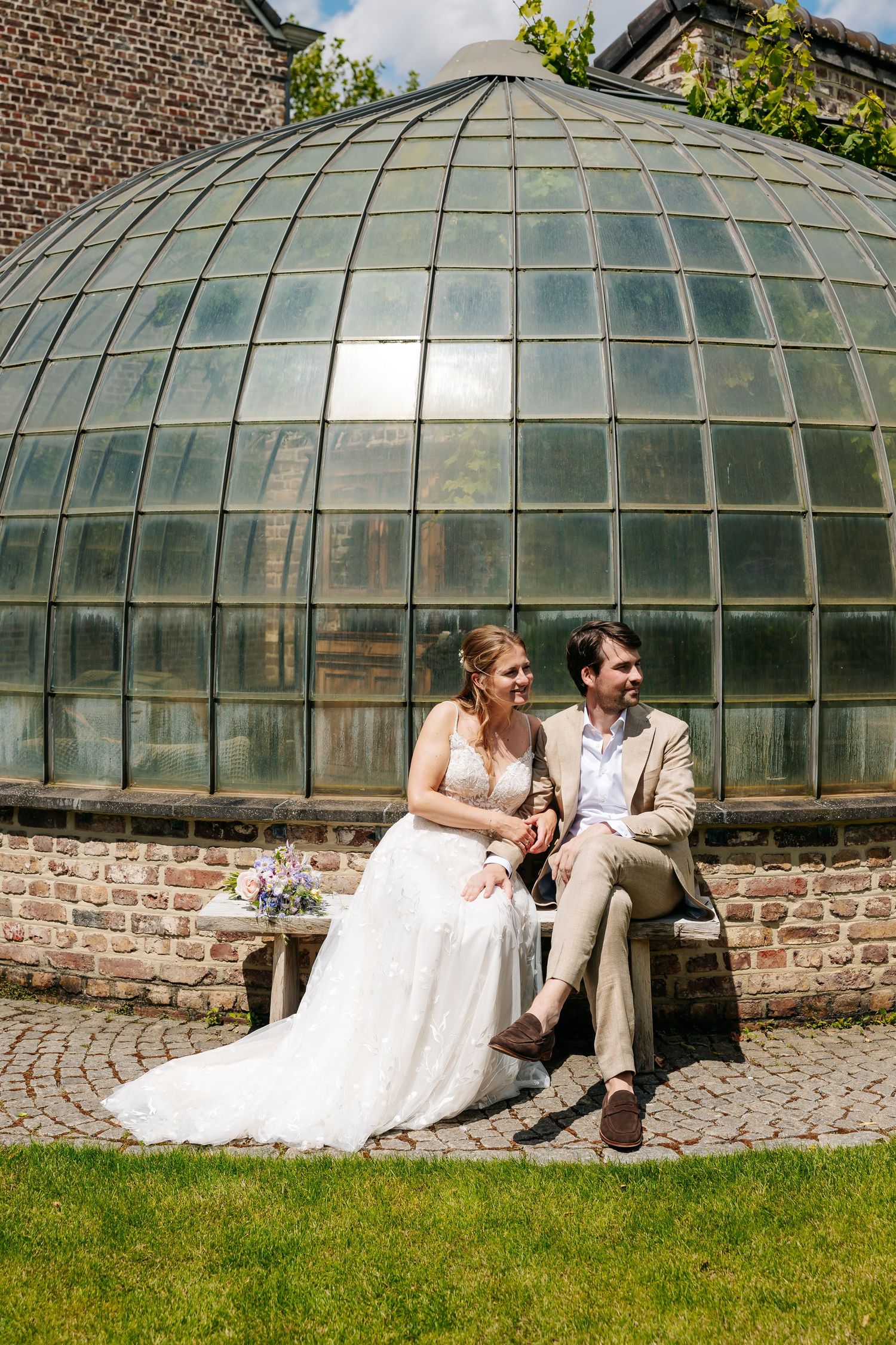Wedding couple seated on a bench before a domed glass greenhouse, bride in white gown and groom in beige suit.