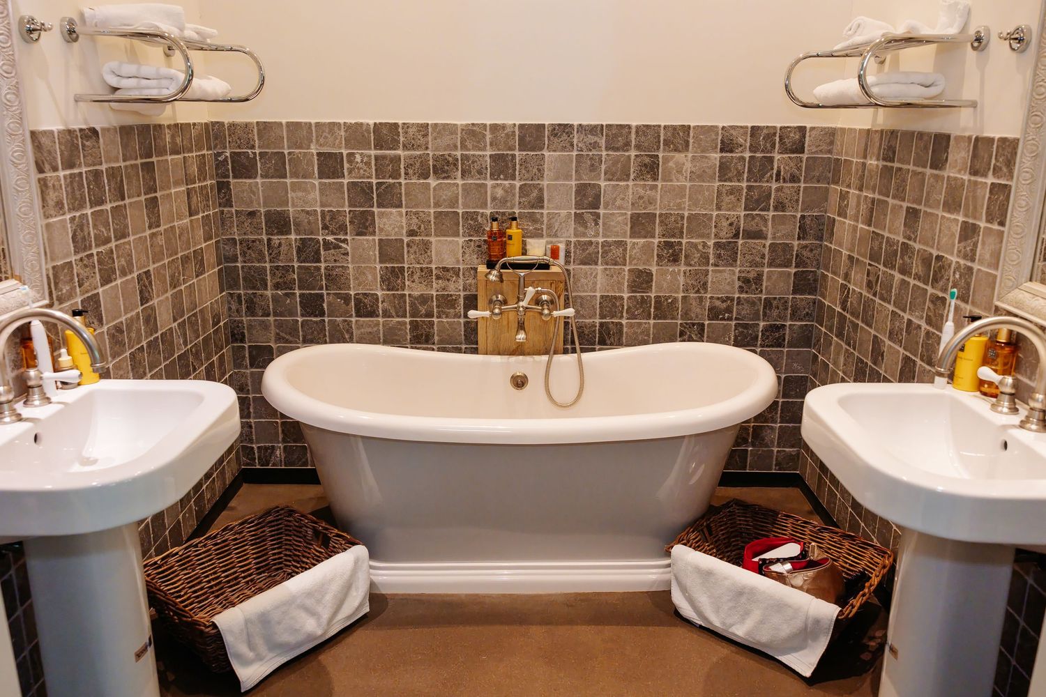 Elegant bathroom featuring a freestanding white slipper bathtub flanked by two pedestal sinks with wicker baskets on tiled floor.