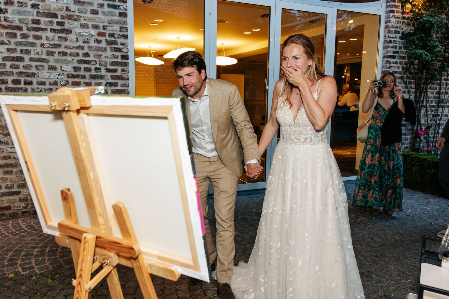 A bride covers her mouth in surprise as she and her groom view a painting on an easel.
