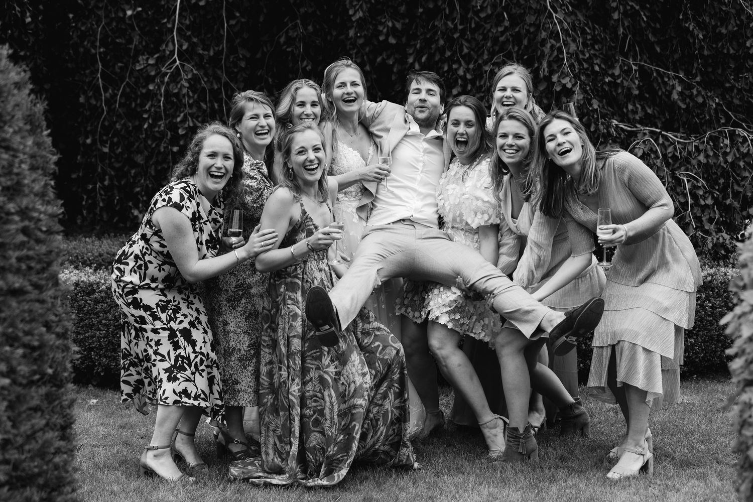 Black and white photo of bride being lifted by laughing bridesmaids in a garden celebration.
