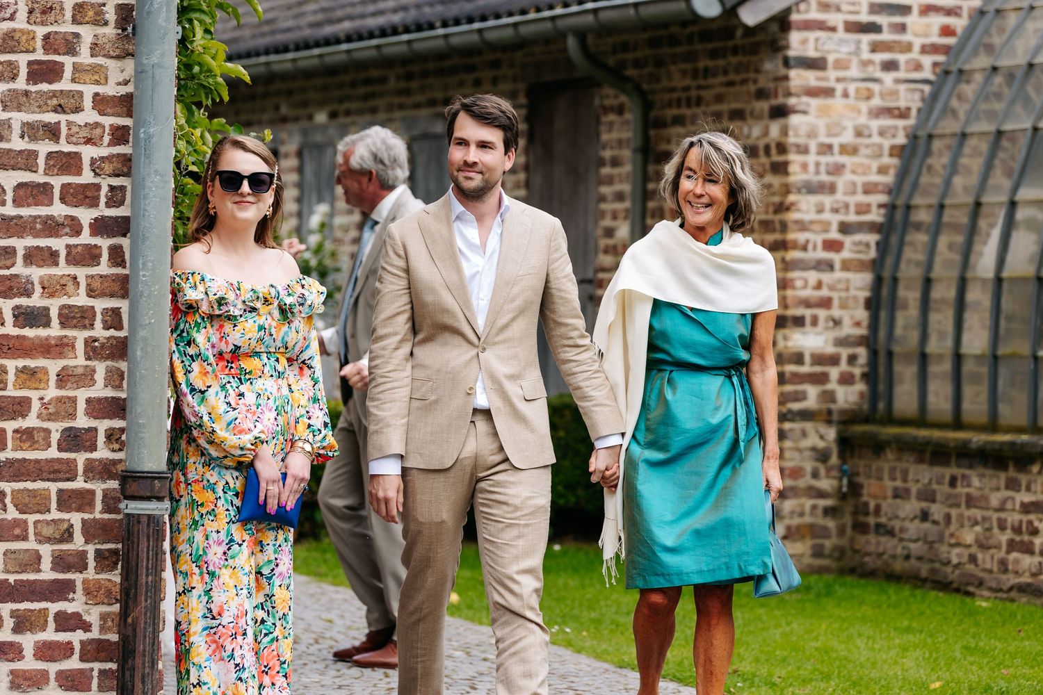 Groom in beige suit walking with two elegantly dressed women at wedding venue.