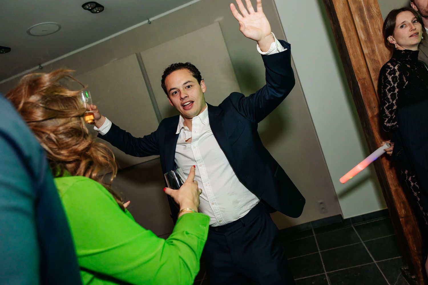 Man in navy suit waves energetically while celebrating at a festive indoor wedding reception.