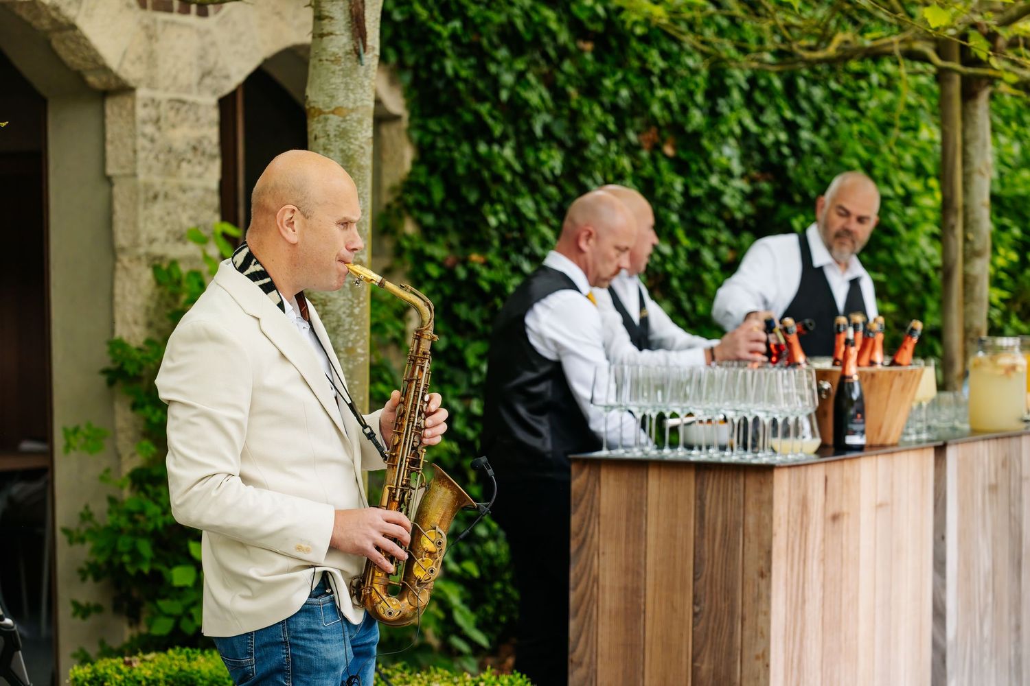 Saxophonist performs in white jacket at outdoor wedding reception while bartenders prepare drinks at wooden bar.