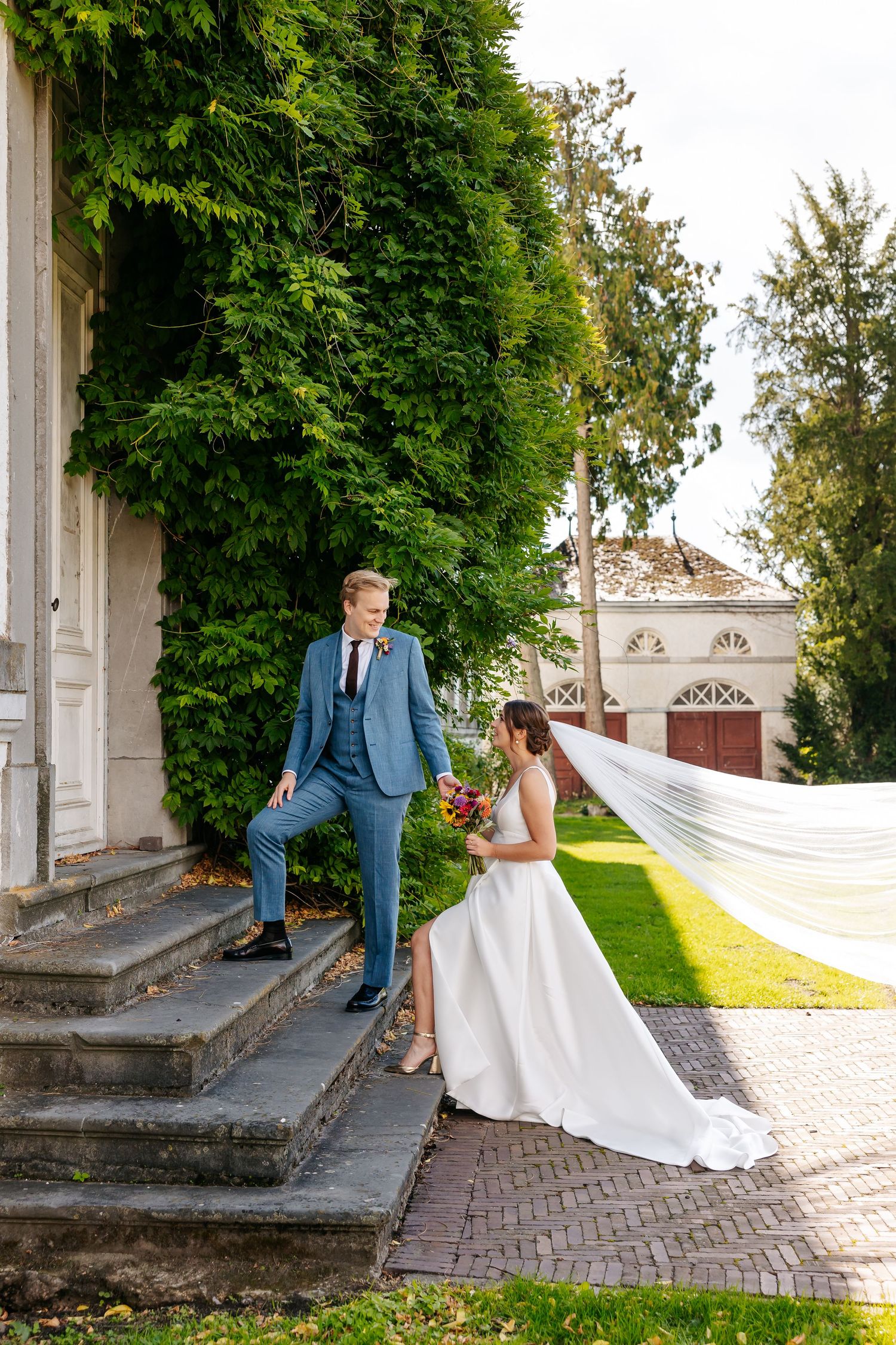 Elegant bride with flowing veil and groom in blue suit posing on stone steps beside ivy-covered building.