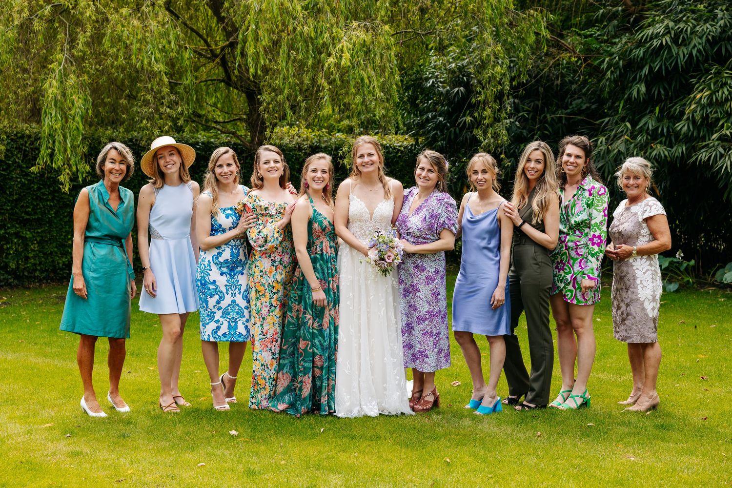 Bride posing with group of female wedding guests on lush green lawn in summer garden.
