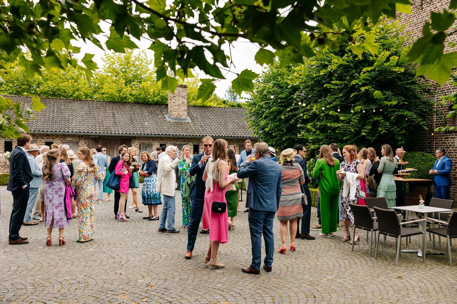 Wedding guests mingle outdoors on a cobblestone courtyard surrounded by greenery.