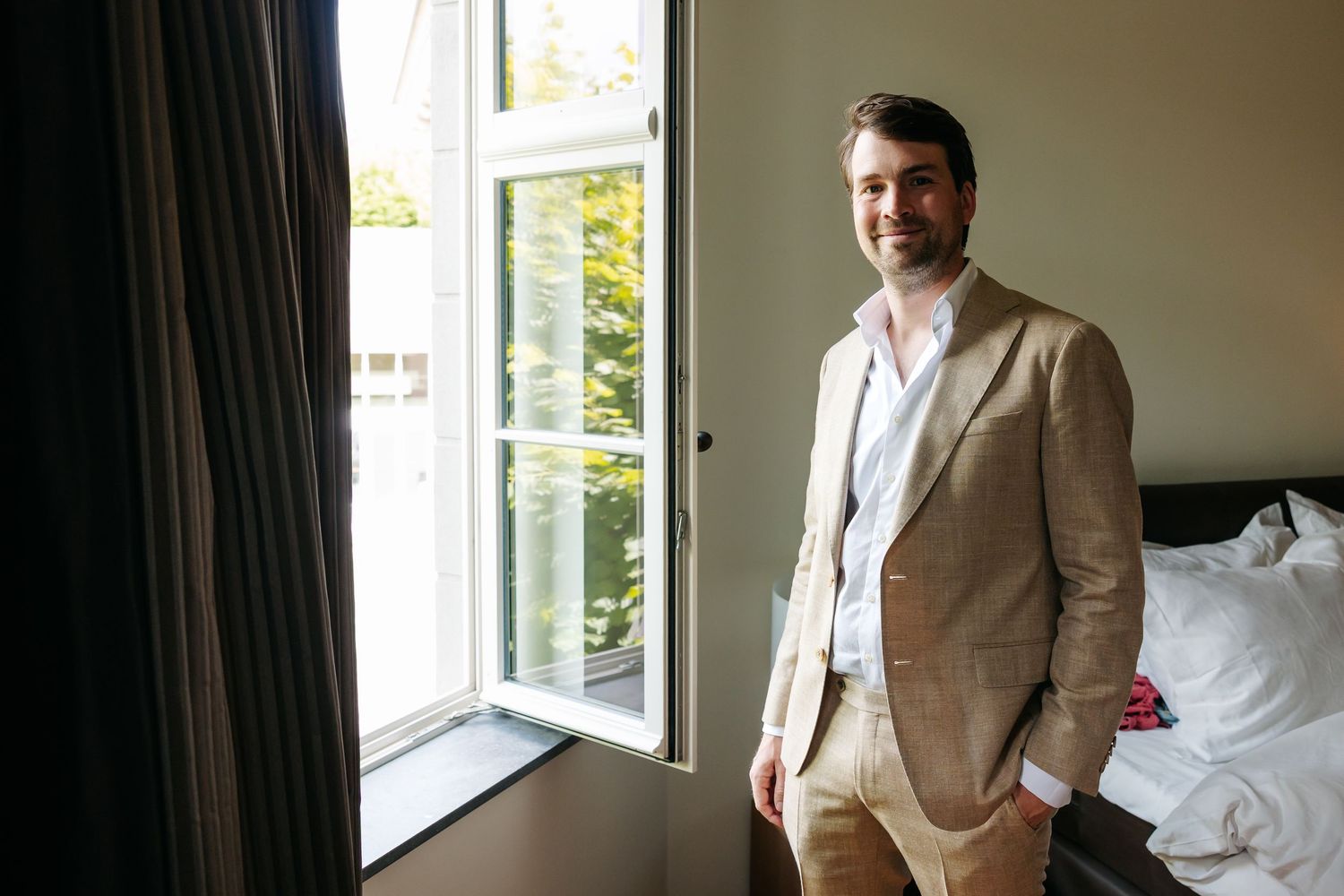 Groom in beige suit standing by window in bright hotel room.