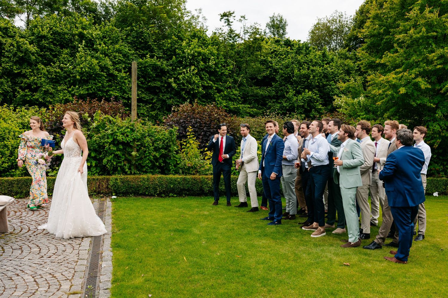 Bride in white gown facing line of wedding guests on lush garden patio during reception.