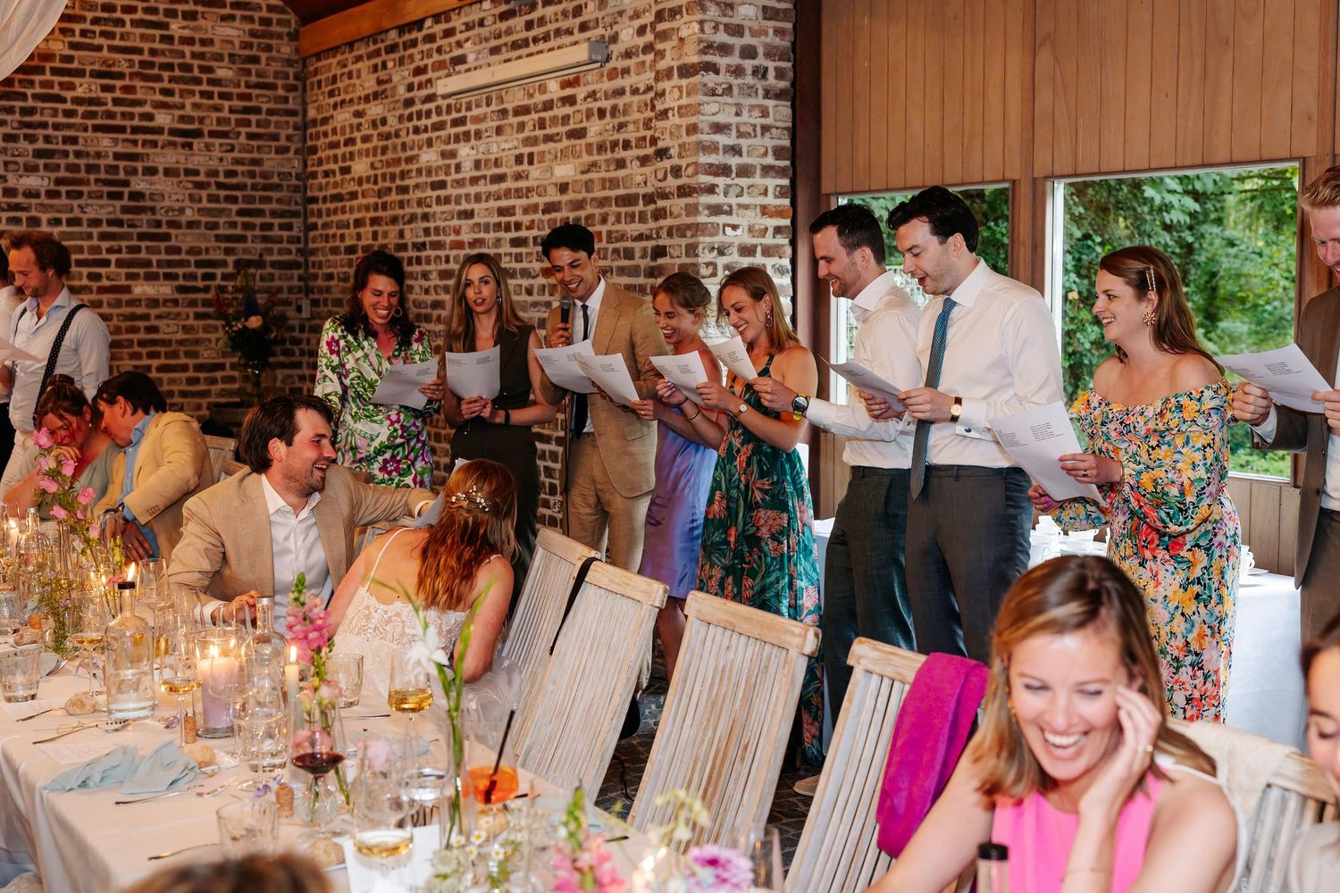 Guests standing and reading from papers during a wedding reception in a rustic brick venue.