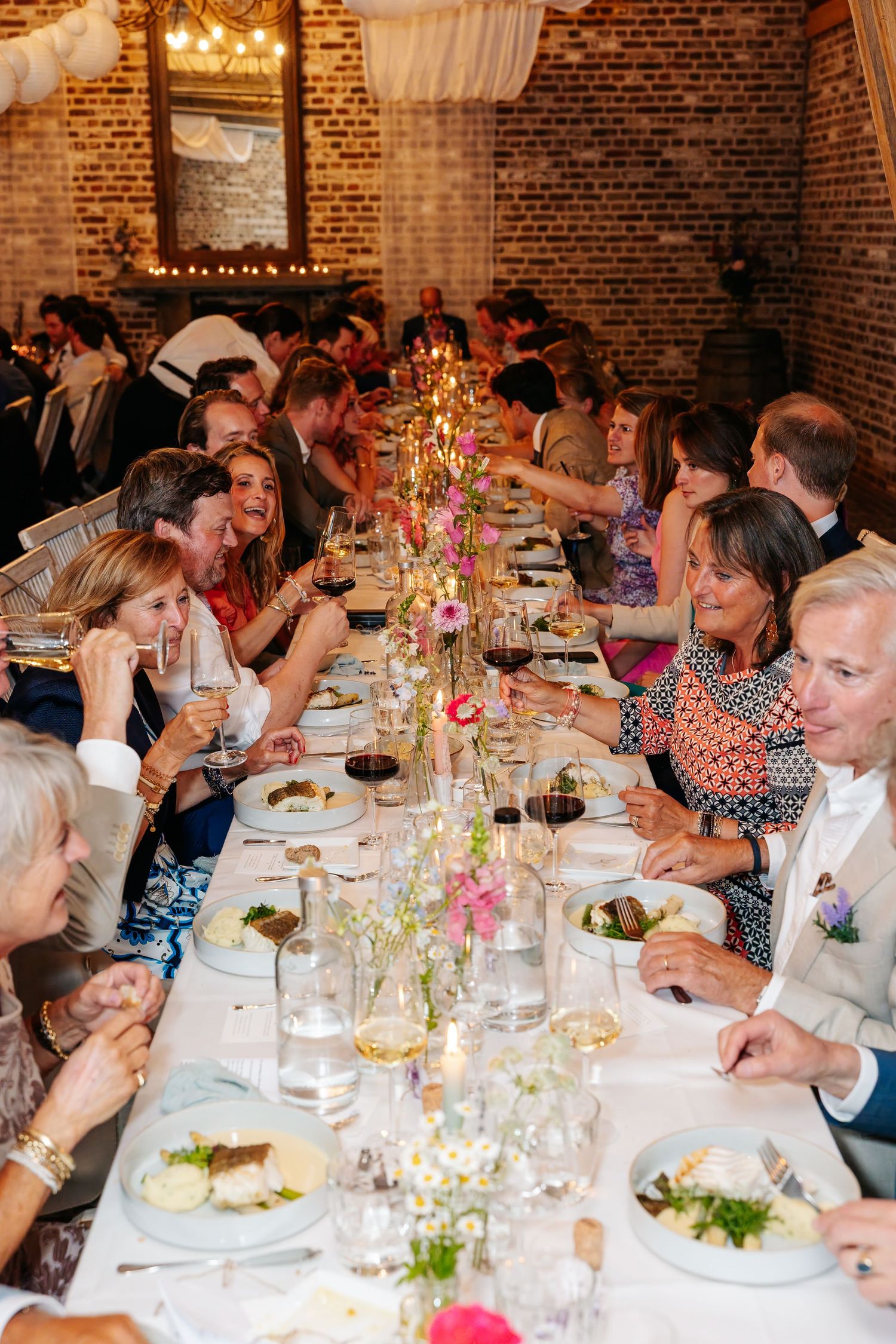 Wedding guests dining at a long decorated table with floral centerpieces in brick venue.