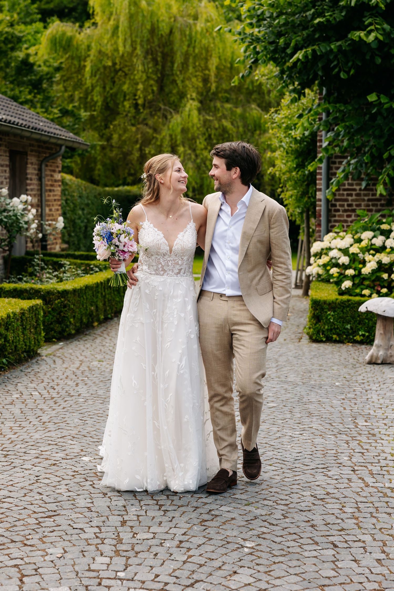 Bride and groom walk hand in hand on cobblestone path through lush garden on their wedding day.