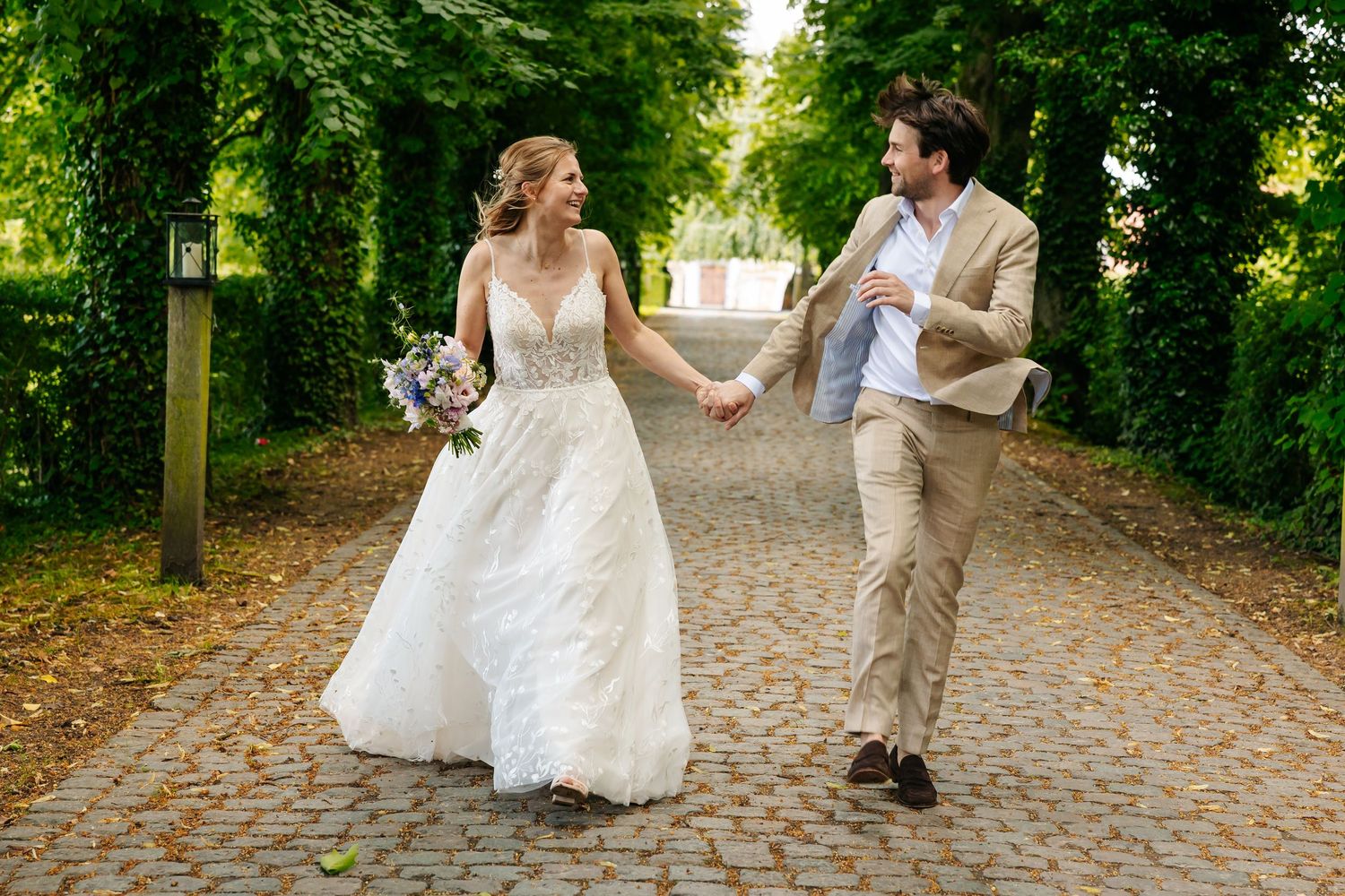 Bride in white gown and groom in tan suit running hand in hand along tree-lined path.
