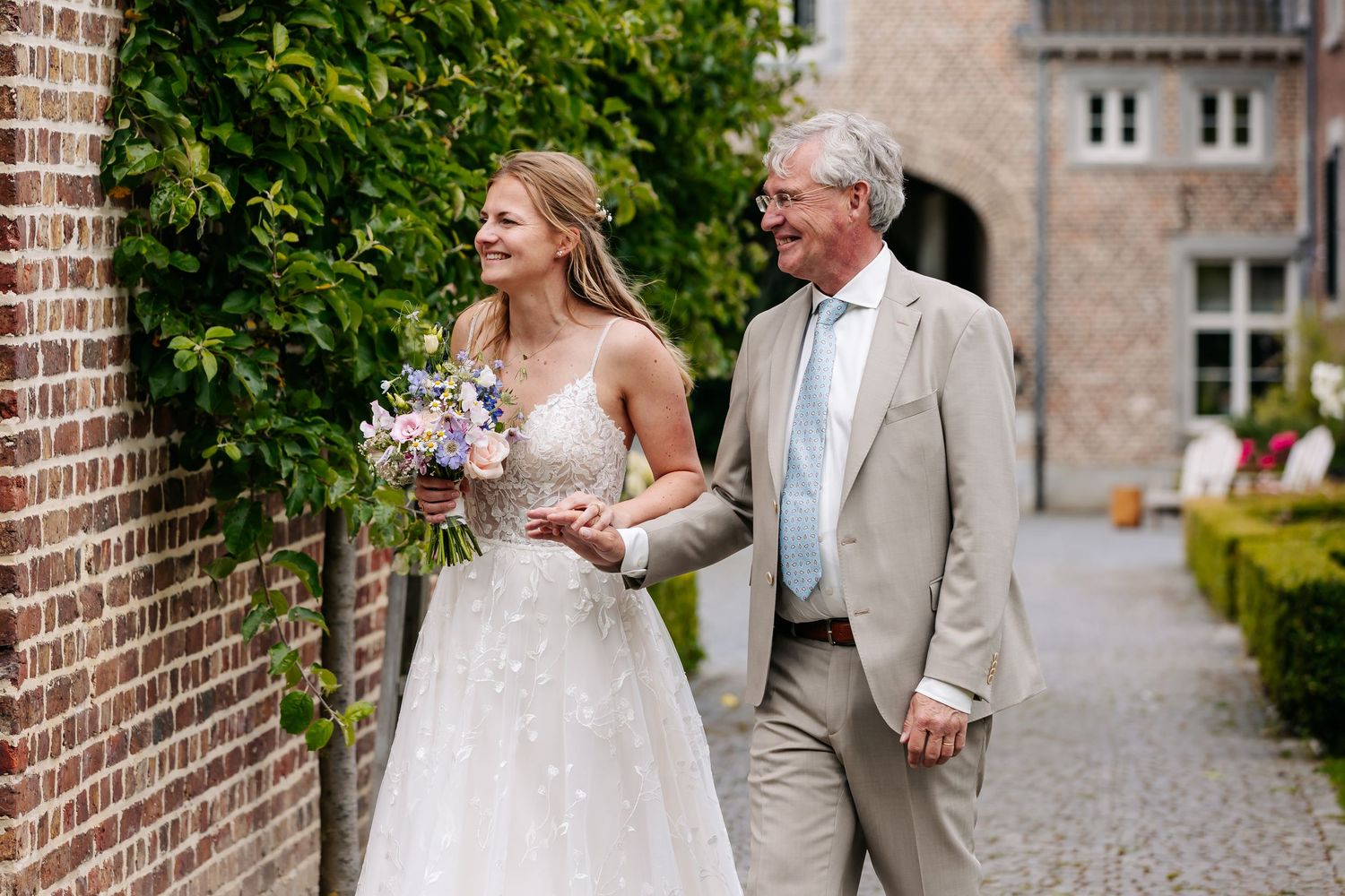 Smiling bride in lace gown holding bouquet walking arm in arm with father outdoors.