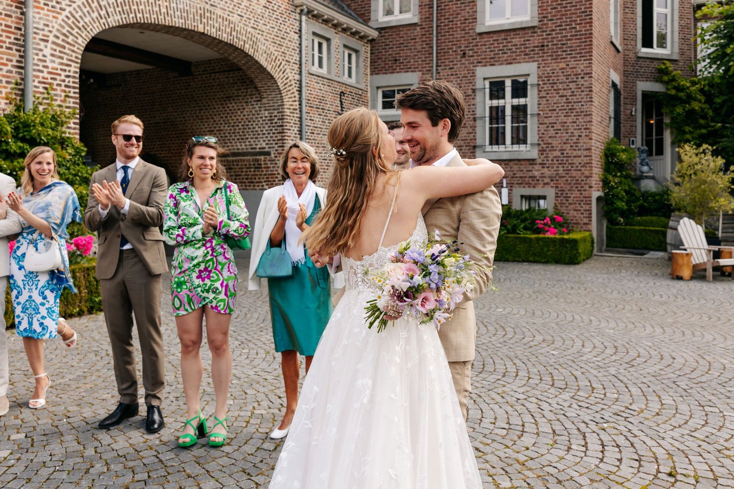 Bride and groom embracing on cobblestone courtyard while wedding guests applaud in background.