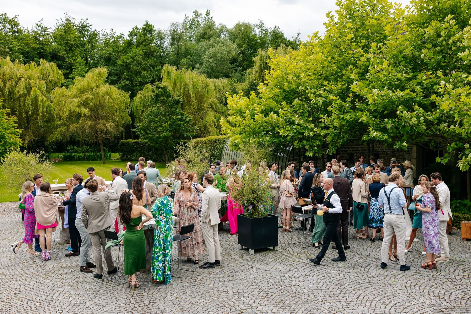 Wedding guests socializing outdoors on cobblestone courtyard surrounded by lush green trees.