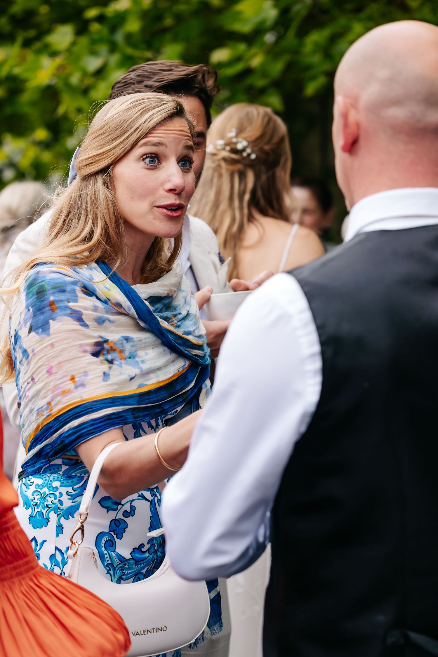 Blonde woman in blue floral dress engages in conversation at an outdoor wedding garden party.