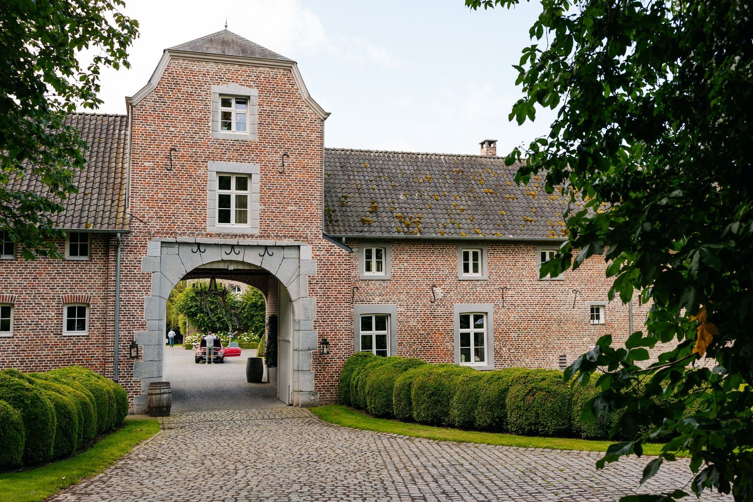 Historic pink brick manor house with arched gateway entrance, cobblestone driveway, and manicured hedges on a sunny day.