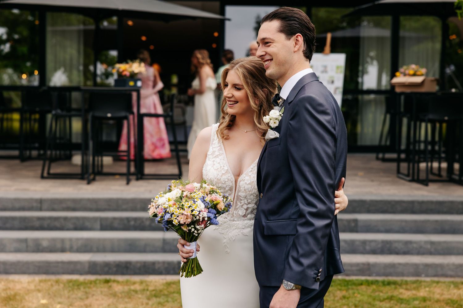 Smiling newlyweds pose together outdoors, bride holding colorful wildflower bouquet in lace wedding dress and groom in navy suit.