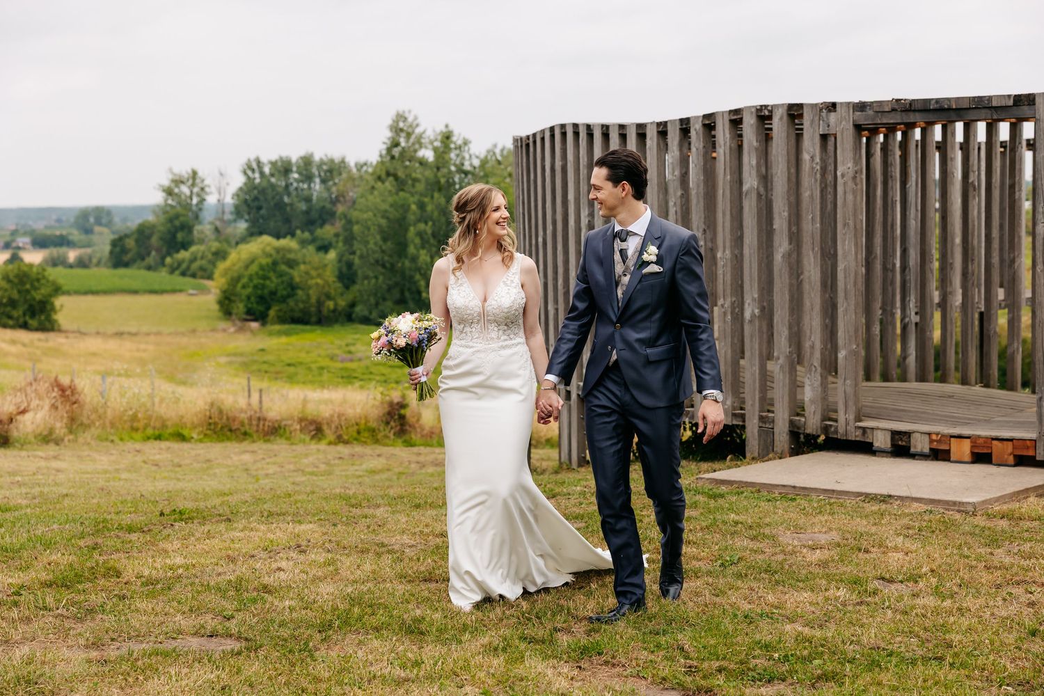 Bride in white gown and groom in navy suit walking hand in hand across green countryside field.
