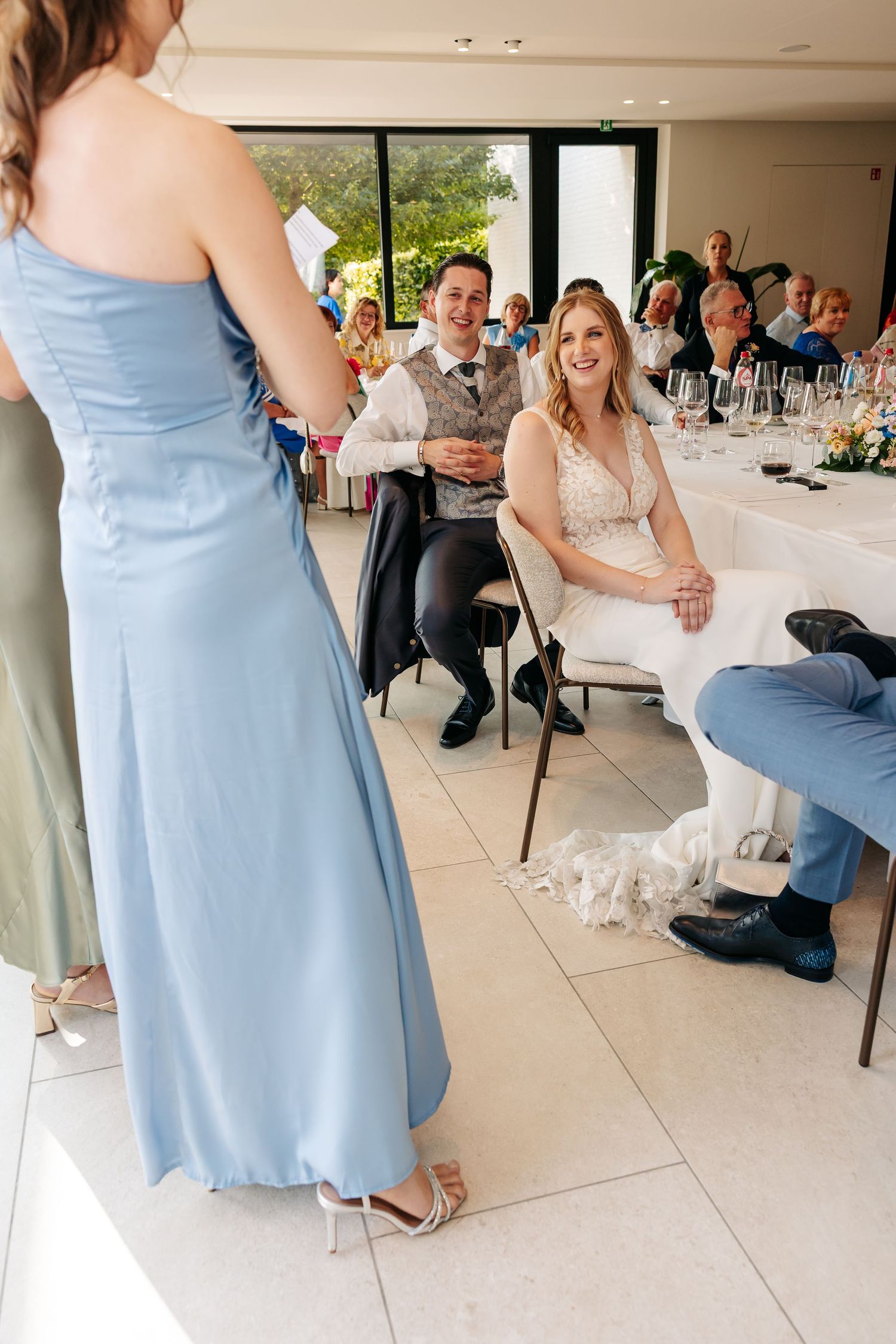 Bridesmaid in blue gown gives speech as bride and groom laugh at wedding reception.
