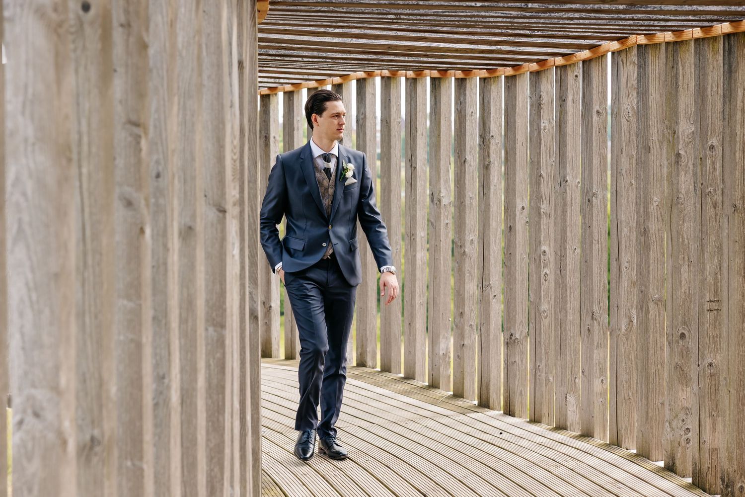 Groom in navy wedding suit walking confidently through a rustic wooden pergola walkway.