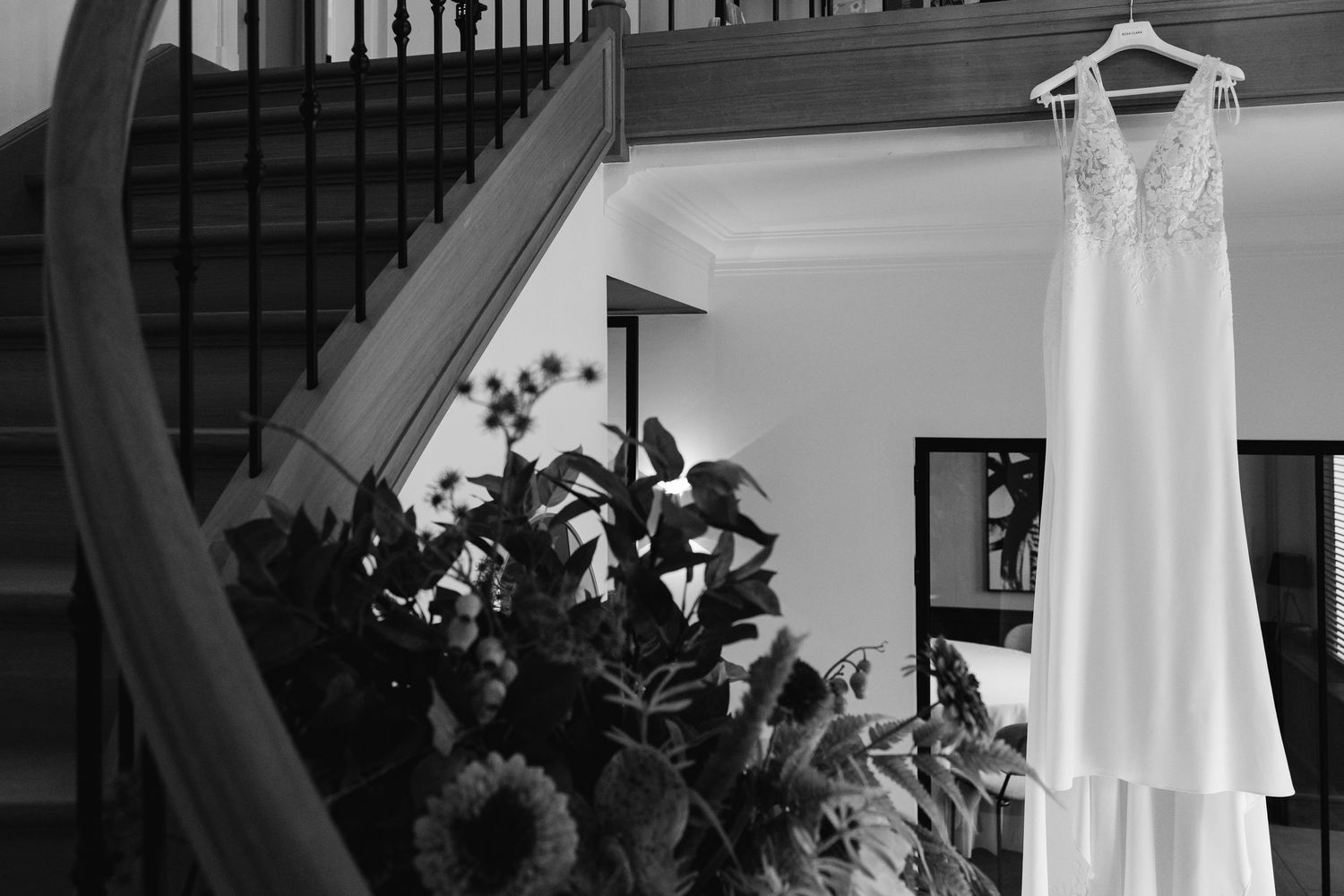 Black and white image of bridal gown hanging near curved staircase with lush floral decor below.