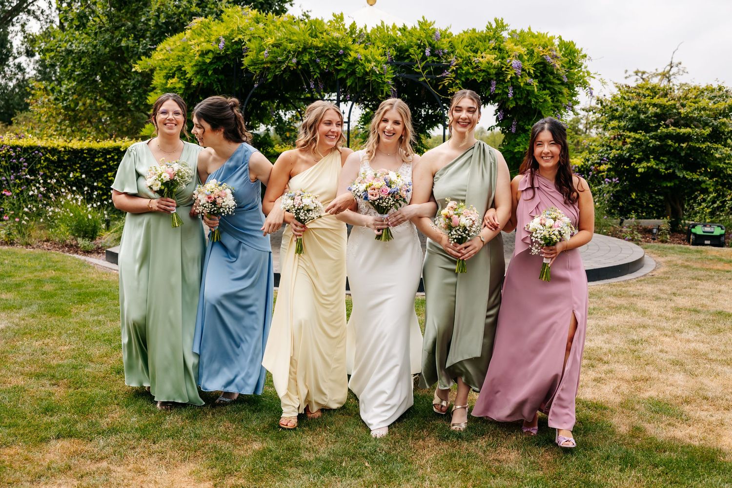 Bride walks with five bridesmaids in mismatched pastel gowns holding floral bouquets in a sunny garden setting.