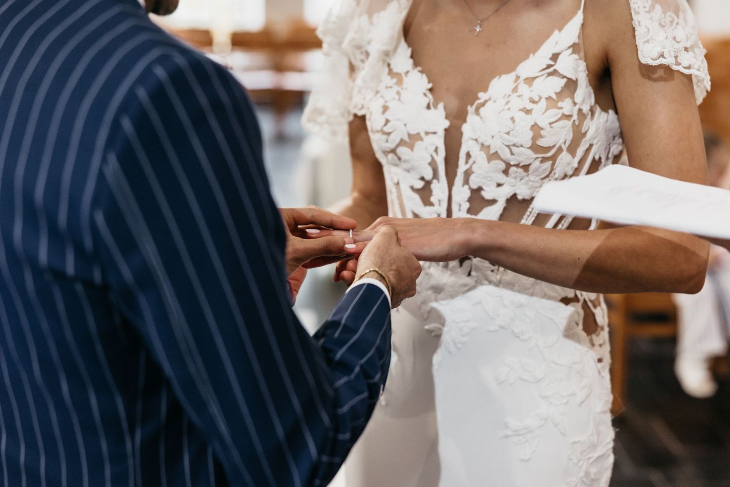 Bride in floral lace gown exchanging rings with groom in navy pinstripe suit at wedding.