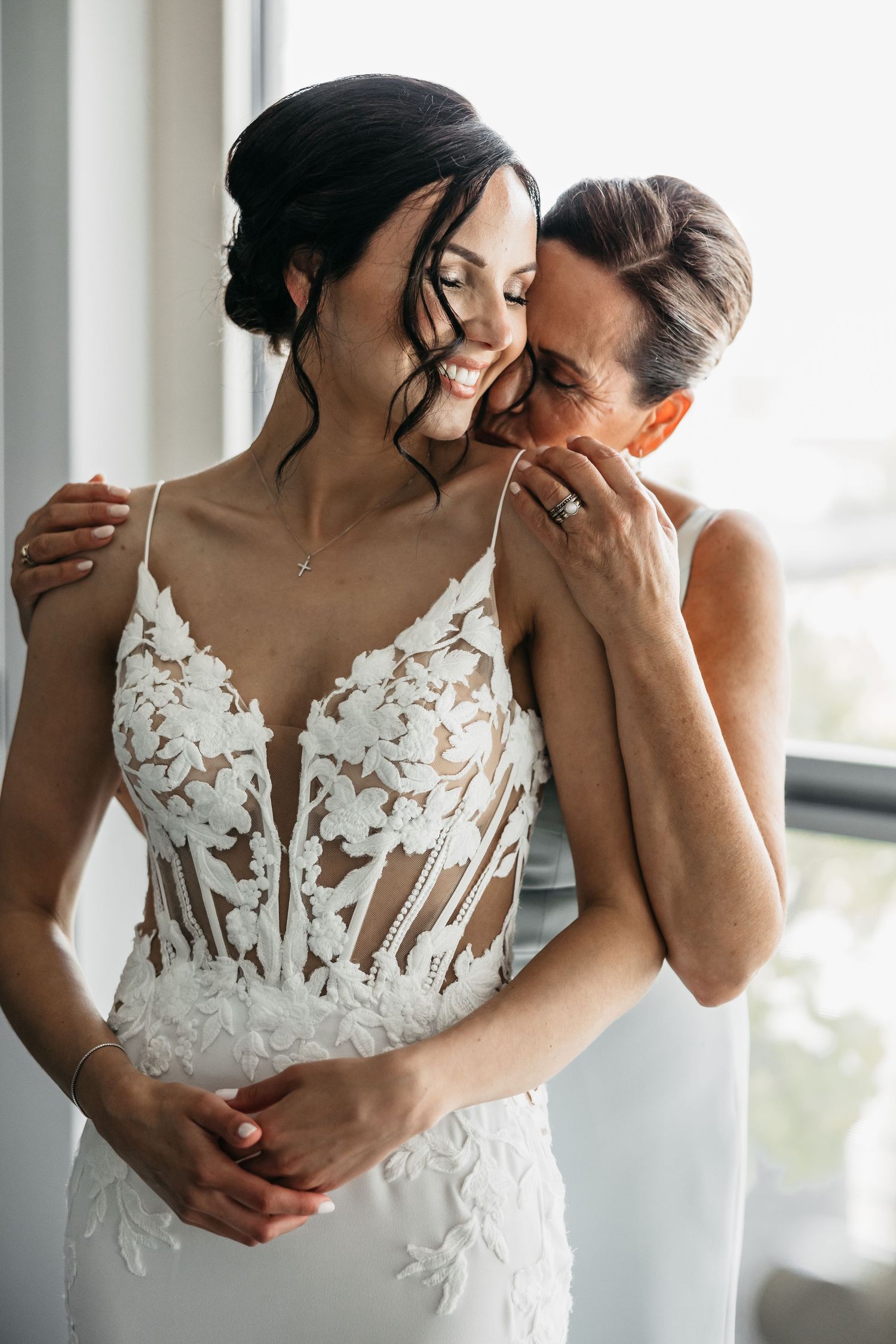 Mother whispering to smiling bride in floral lace wedding gown by window.