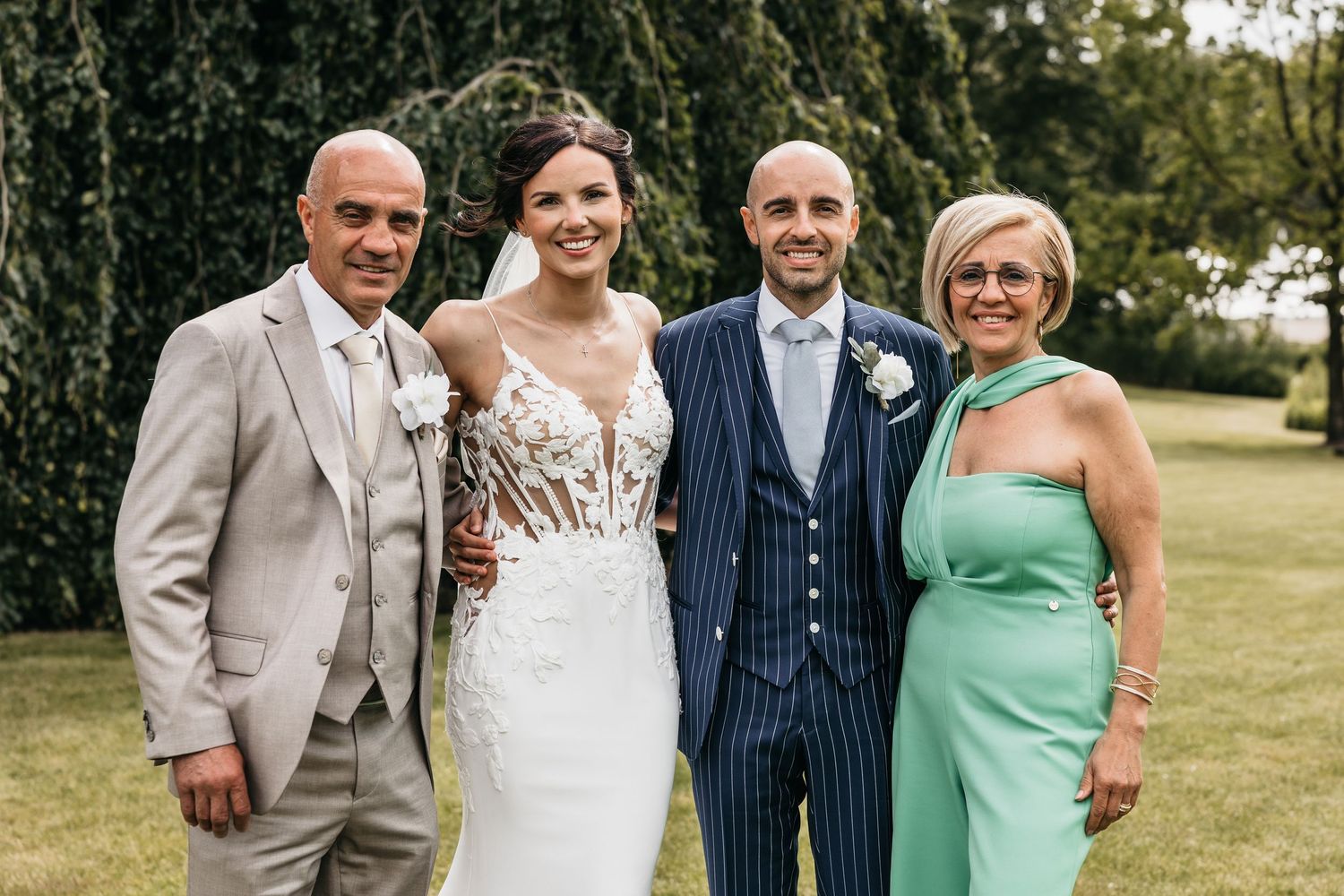 Wedding couple posing with family outdoors, bride in lace gown, groom in navy pinstripe suit.