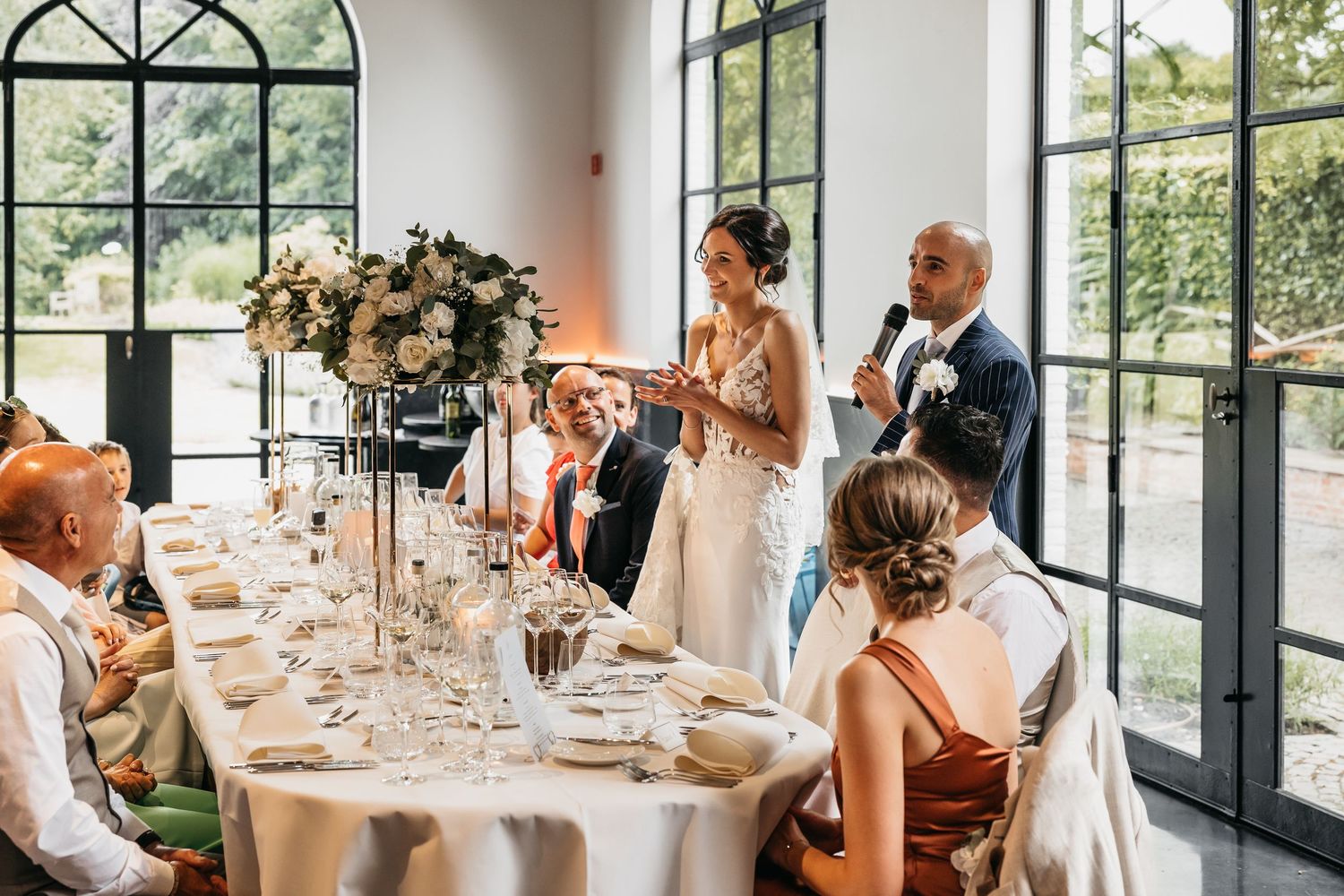 Bride and groom give heartfelt speech at elegant wedding reception table adorned with tall floral arrangements.