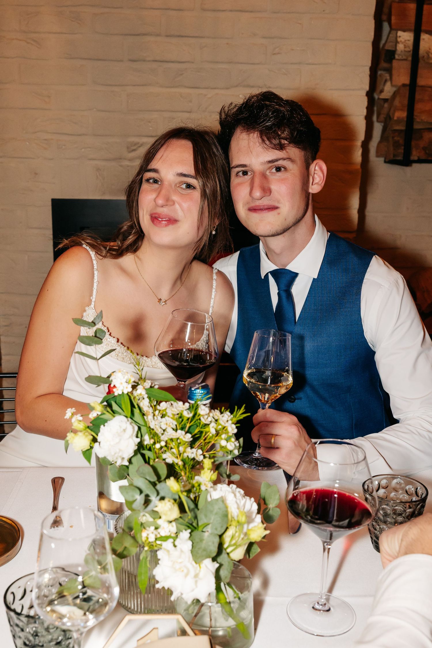 Young couple holding wine glasses and smiling at wedding reception with floral centerpiece.