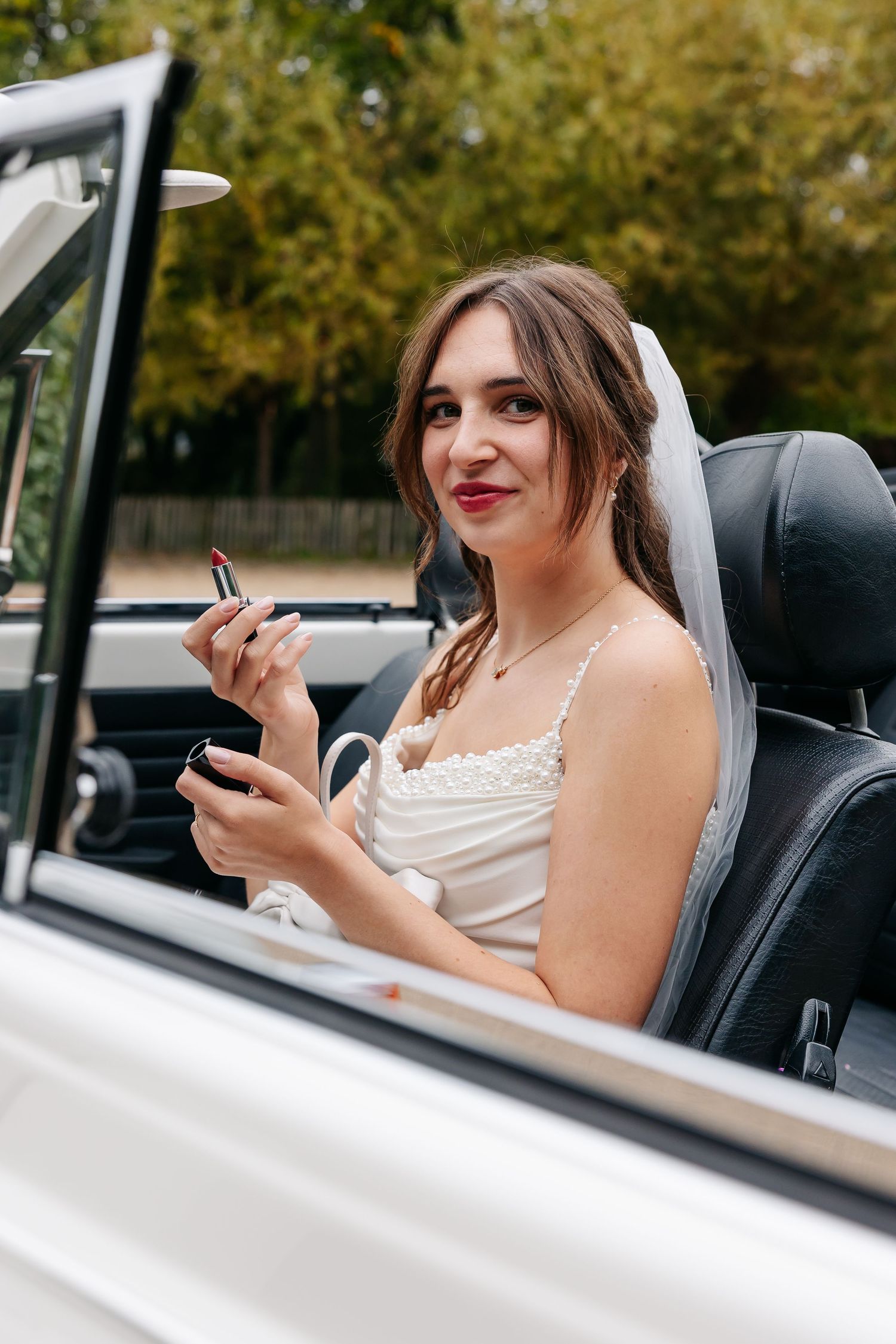 Bride in white gown and veil sits in white convertible car applying red lipstick using a compact mirror.